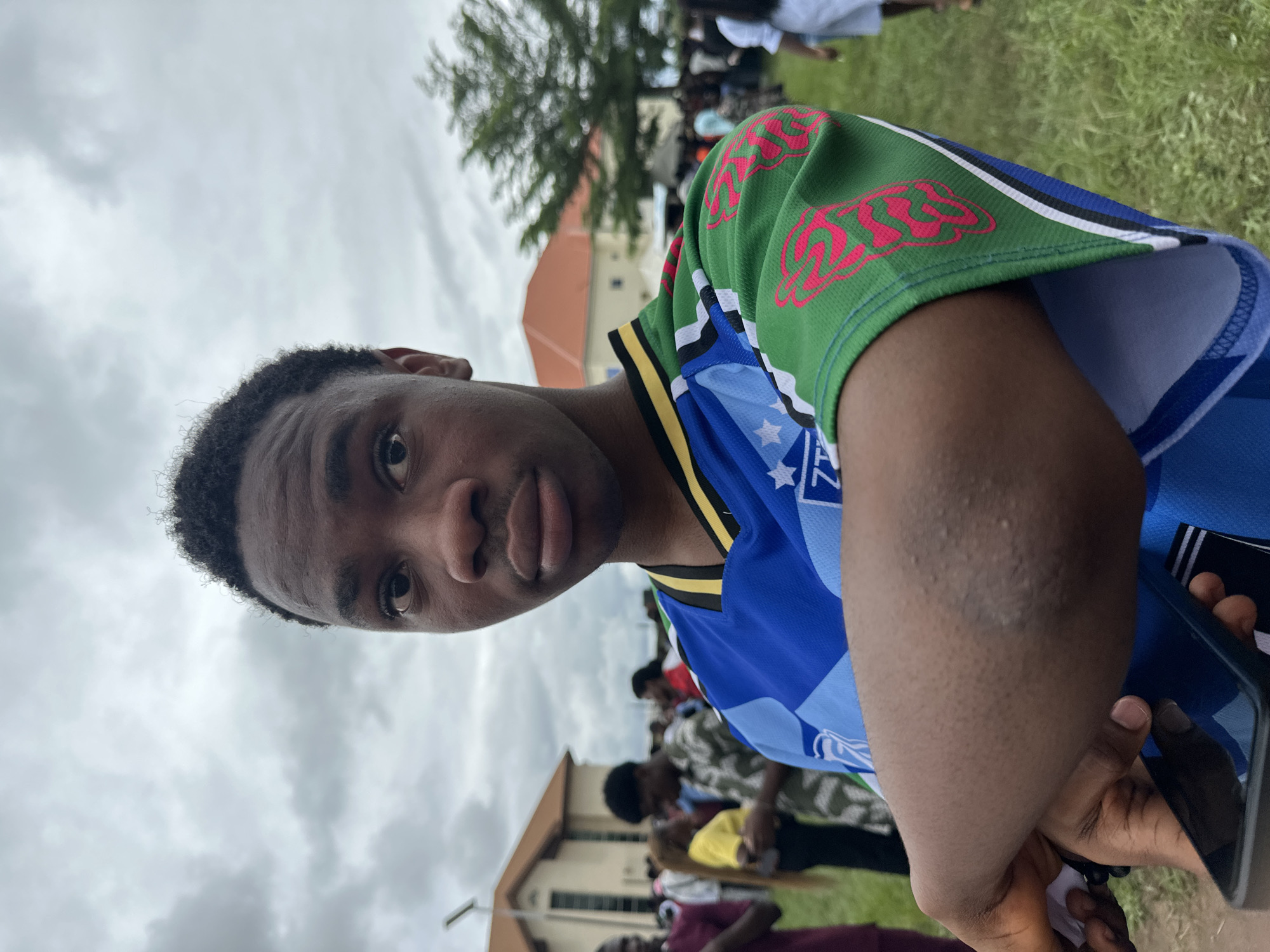A medium shot captures a young Black male from the chest up, standing outdoors. He has dark, short, curly hair and is looking directly at the camera with a neutral expression. He is wearing a blue and green jersey with white and yellow trim. The jersey has a pattern of blue and white squares on the front, and the sleeves are green with a pink graphic design. His left arm is crossed over his chest, and he is holding a dark-colored smartphone in his right hand.
The background suggests an outdoor event or gathering. To the left, a light-colored building with an orange roof is visible. In the mid-ground, a crowd of people can be seen, some blurred due to the shallow depth of field. Green grass covers the ground. The sky is overcast, with gray clouds. The location is identified as Ihiagwa, Nigeria.
