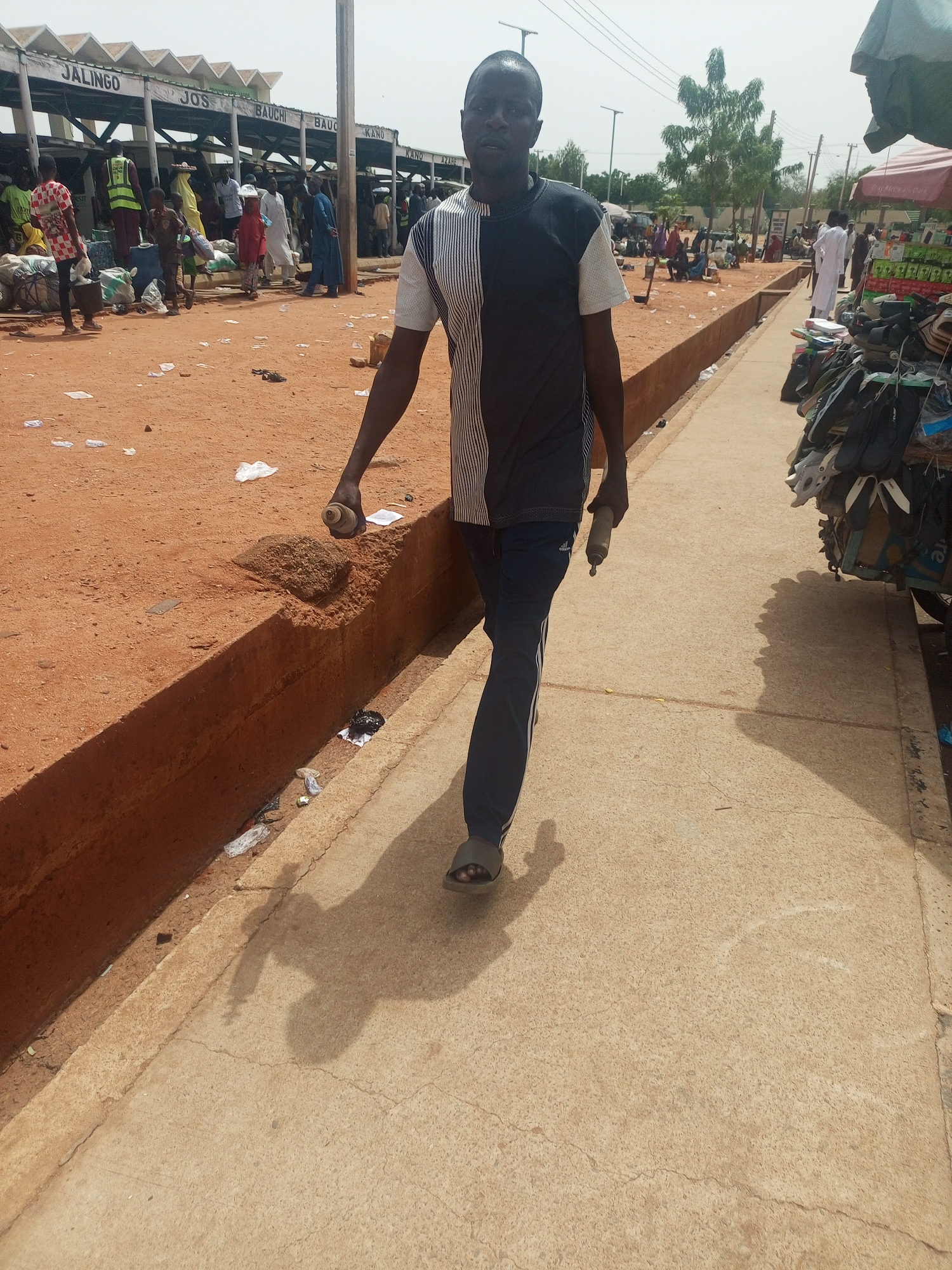 This image depicts a bustling outdoor market scene in Gombe, Nigeria, under bright daylight. A man, who is the primary subject, walks on a paved concrete path towards the viewer. He wears a black and white paneled short-sleeved shirt, dark athletic trousers with white stripes, and sandals. He holds a light-colored cylindrical object in his left hand and a darker one in his right, possibly small spray cans or bottles. His shadow is distinctly cast on the ground to his left.
To the man's left, a vibrant market area unfolds on reddish-brown dirt ground, visibly strewn with litter including plastic bags and wrappers. A low concrete barrier delineates the edge of the paved path from this sandy terrain. In the background, numerous people are engaged in market activities, moving around or congregated near various stalls. Dominating this area are several large, open structures with corrugated metal roofs, identified as market sections or transport points by signs bearing names of Nigerian cities: "JALINGO," "JOS," "BAUCHI," "BAUO," "KANO," and "AZABE." Shoppers and vendors, some carrying bundles of goods, are dressed in a mix of traditional and casual attire.
On the right side of the paved path, a vendor's stall is partially visible, displaying an assortment of shoes and sandals hanging from racks. A small red tent is also present further down the path. The scene conveys the lively and somewhat dusty atmosphere of a busy local market, characterized by clear skies and strong sunlight.