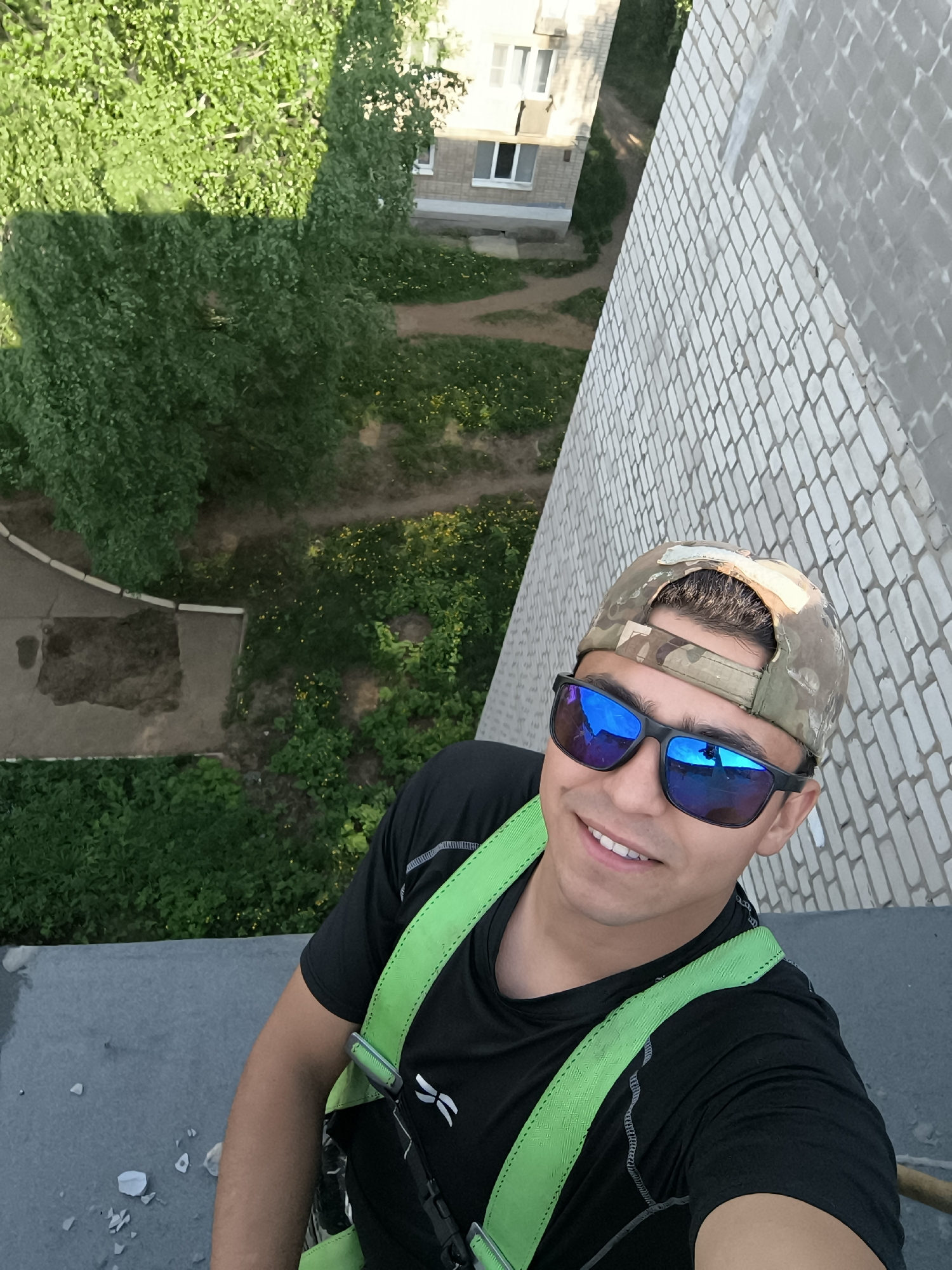 The photograph is taken from an elevated perspective, looking down at a man. He is wearing a black short-sleeved shirt, a camouflage baseball cap, and sunglasses. A green harness is strapped around his upper body. The man is smiling at the camera.
The background includes a white brick wall adjacent to the man, and trees and a building with windows in the distance. The photo's location is in a city of Unknown, Unknown.
