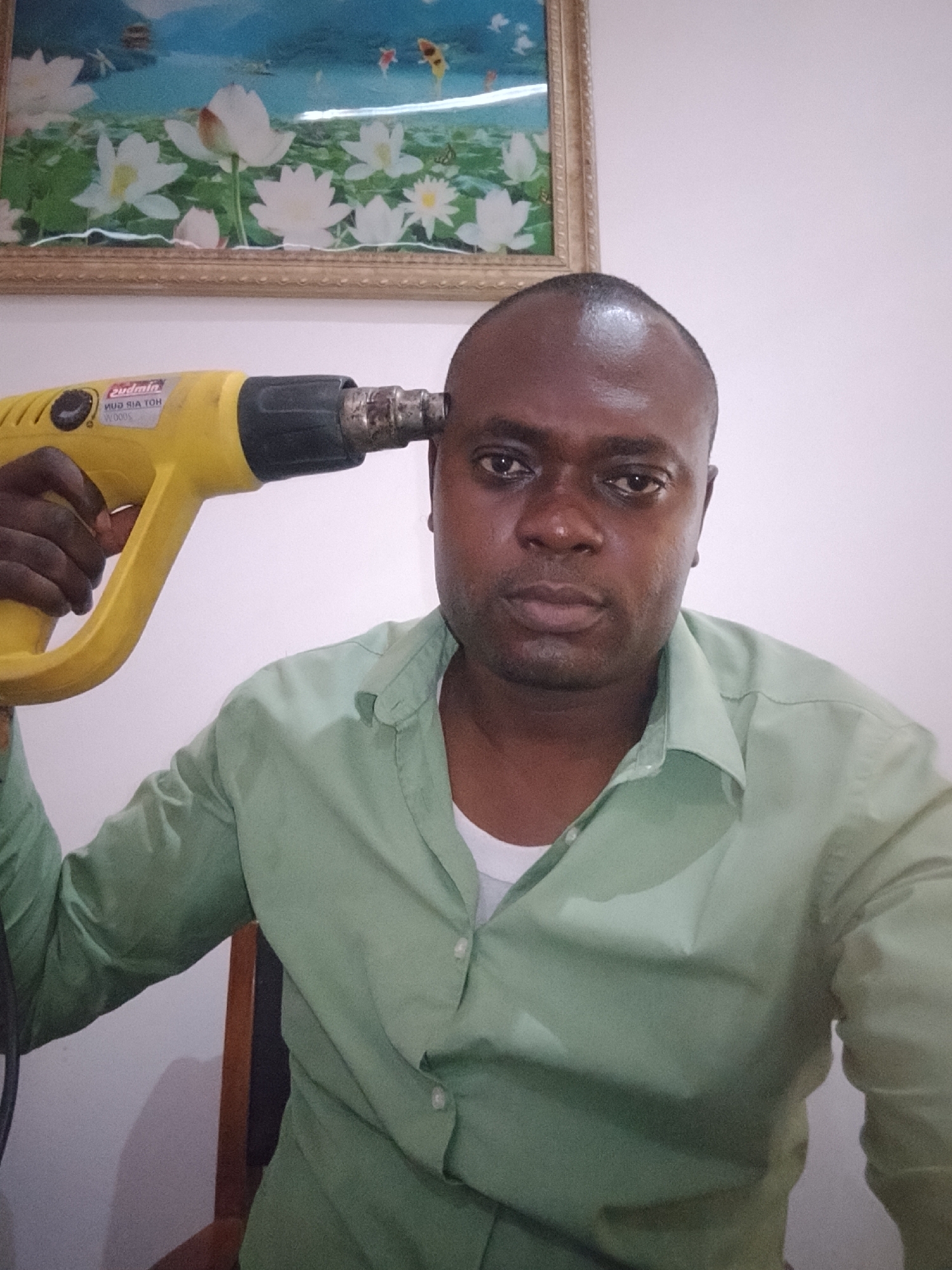 A man with dark skin and short black hair, wearing a light green button-down shirt over a white undershirt, is sitting on a wooden chair in front of a white wall. He is holding a yellow and black heat gun against his head. Behind him, there is a framed picture of white lotus flowers and blue water. This picture was likely taken indoors in Gwarinpa, Nigeria.