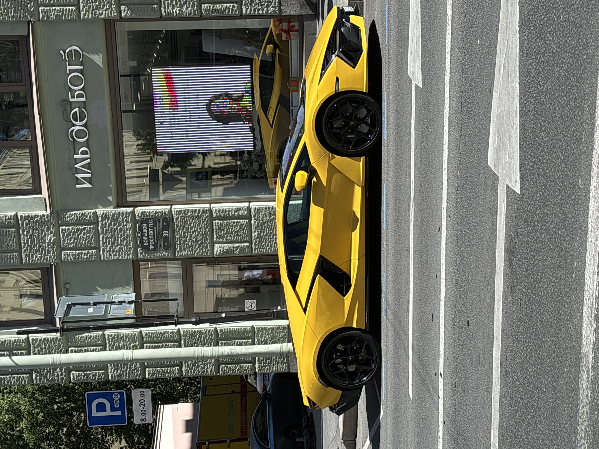 A bright yellow Lamborghini Aventador is prominently featured on a city street, positioned parallel to the curb on the right side of an asphalt road. The car has black multi-spoke wheels.
In the background, a multi-story building with a textured, light greenish-gray stone facade is visible. The ground floor features large display windows. Above the windows, a prominent store sign in Cyrillic reads "ИЛЬ дЕ БОТЭ" (IL DE BEAUTE), indicating a cosmetics and perfume retailer. To the left of the store sign, a street sign identifies the location as "БОЛЬШОЙ ПРОСПЕКТ П.С." (Bolshoy Prospekt Petrogradskaya Storona), with the building number "39" clearly displayed, along with adjacent numbers "41" and "37".
Further left, a blue square parking sign with a white "P" and numbers "10 15 20" is mounted on a pole. Below it, a white rectangular sign indicates parking hours as "8.00 - 20.00". A large green tree is partially visible behind the parking sign.
Reflections in the shop windows show a pedestrian crossing sign, a distorted image of a person, and a yellow car, likely a reflection of the Lamborghini itself. Inside the store, a vertical LED display with colorful abstract patterns is visible. Red gift-wrapped boxes are also reflected within the display window.
Behind the Lamborghini, a dark-colored sedan is partially visible parked on the sidewalk. Further back and to the left, a yellow utility truck with "ВОДОКАНАЛ" (VODOKANAL - water utility) written in Cyrillic is parked.
The asphalt road surface features clear white painted lines, including solid lines marking lanes and white arrow markings indicating traffic direction. The scene is illuminated by bright daylight, casting distinct shadows.
The location is identified as Bolshoy Prospekt Petrogradskaya Storona, number 39, in the city of Saint Petersburg, Russia.