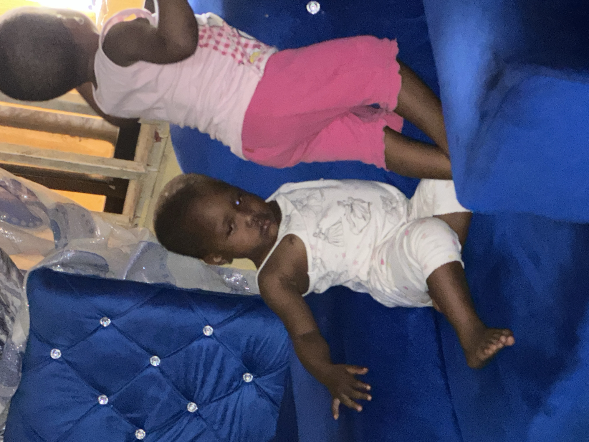 The image captures two young children on a bright blue, tufted sofa. The child in the foreground, facing the camera, is a young Black girl wearing a white onesie with faint drawings of princesses. She is sitting with her legs crossed, her left arm extended to the side and her right hand resting on her lap. Her expression appears weary or sleepy. Behind her, to the right, stands another young Black child, also a girl, facing away from the camera. She is wearing a pink tank top with a checkered pattern and bright pink shorts. The setting appears to be indoors, with a window and possibly some furniture visible in the background. The overall lighting suggests it may be daytime. There are no indications of the city of Narayi, Nigeria within the image itself.