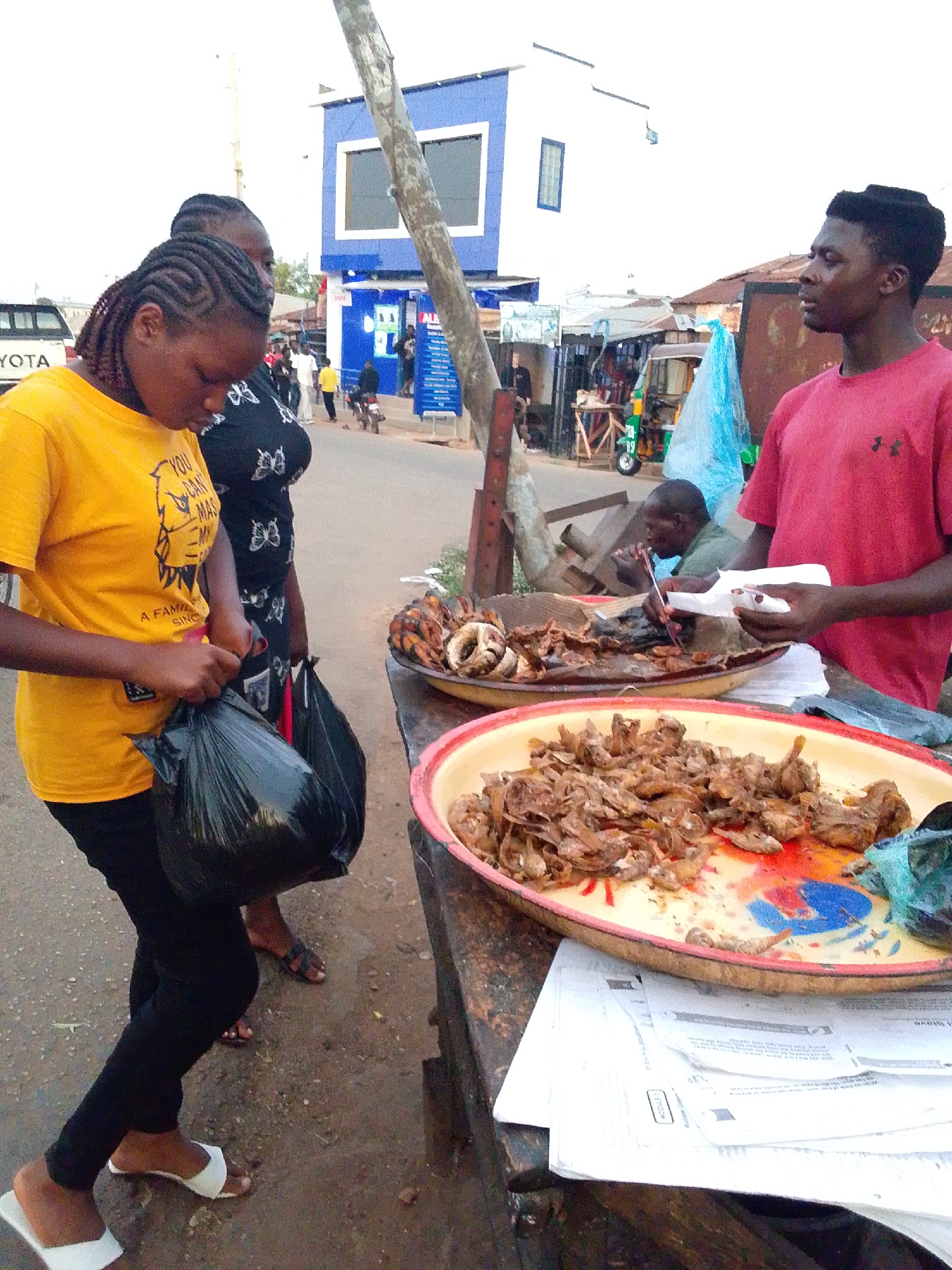 Here's a detailed description of the image:
**Overview**
The image captures a street scene in Jos, Nigeria, focusing on a street food vendor and two female customers. The setting appears to be an outdoor market or roadside stall.
**People**
* **Vendor:** A young man stands behind a table displaying food. He wears a red t-shirt and is holding a piece of paper. He appears to be interacting with the customers.
* **Customers:** Two women are looking at the food. One woman wears a yellow t-shirt, black leggings, and white slides. She is putting something into a black plastic bag. The second woman, standing behind her, wears a black top with butterfly patterns and dark sandals.
* **Other People:** There are several other people visible in the background, walking or standing near buildings across the street. Another man is visible sitting at the vendor's stall, appearing to be eating.
**Subjects & Objects**
* **Food Display:** There are two large trays of fried food on the table. One tray contains fish, and the other contains what appears to be fried chicken parts.
* **Table/Stall:** The trays are placed on a weathered wooden table covered with newspaper at the bottom.
* **Bags:** The woman in the yellow shirt is holding a black plastic shopping bag.
* **Environment:** The background includes a blue and white building, other street vendors, a motorcycle taxi (keke), and other shops. The ground appears to be paved with a mix of asphalt and dirt. A wooden pole stands near the table.
**Actions & Interactions**
* **Purchasing Food:** The customers seem to be in the process of buying food from the vendor. The woman in yellow is preparing to place something into her bag, indicating she has made a purchase.
* **Vendor Serving:** The vendor appears to be holding a piece of paper for packaging or wrapping the food.
**Environment**
* The scene takes place outdoors, likely on a street or market area.
* The lighting suggests it is daytime.
* The overall atmosphere is busy and indicative of a bustling commercial area.