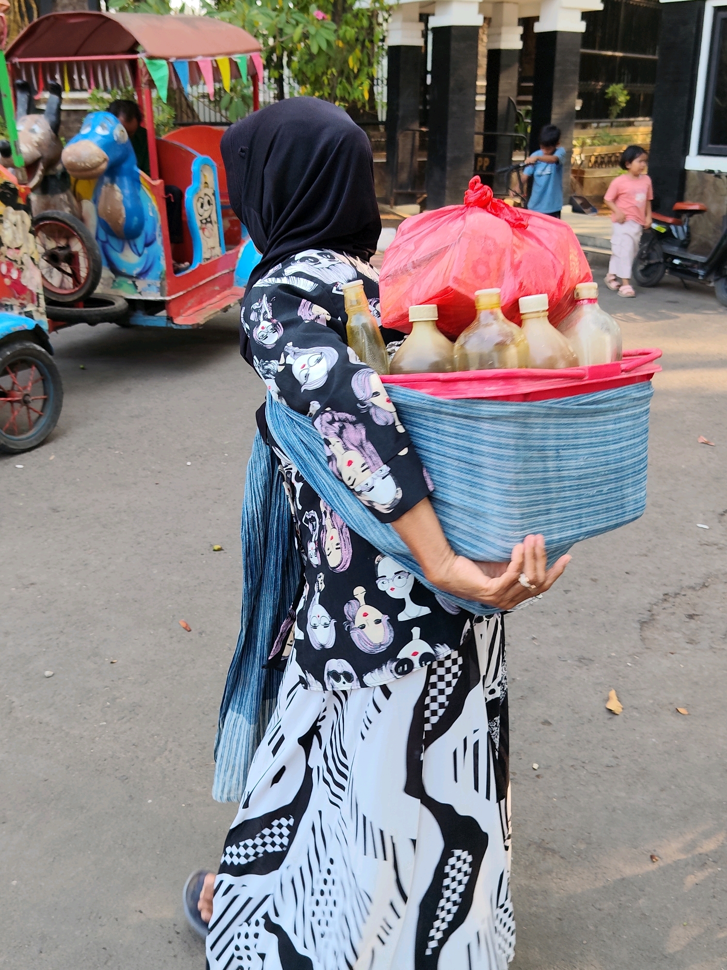 A woman wearing a black hijab, a black and white patterned shirt with cartoonish faces, and a white and black patterned skirt walks to the right, carrying a blue striped basket filled with bottled drinks and a large red plastic bag. The basket is draped with a blue scarf. In the background, a red and blue toy train is parked. Two children are visible further in the background, one in a blue shirt and another in a pink shirt. The scene takes place on an asphalt road in Purwakarta, Indonesia, with buildings and greenery in the background.