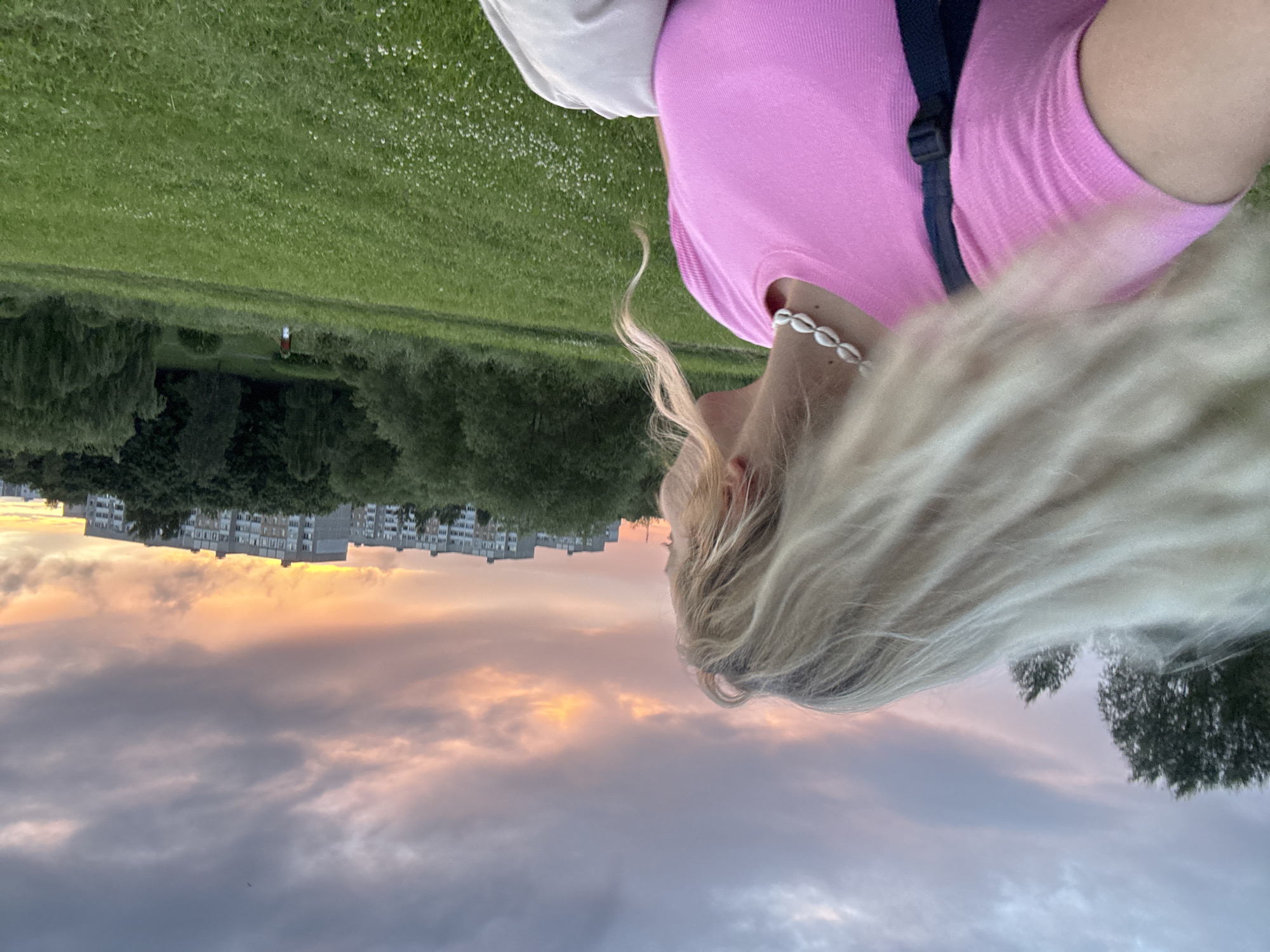 The image shows a young Caucasian woman with long, blonde hair, wearing a pink top and a seashell necklace, standing in a grassy area. Her hair is being blown by the wind to her left. In the background, there are trees, apartment buildings, and a sunset sky with clouds. The location is identified as the park area near the Minsk National Library in Minsk, Belarus. The time appears to be late afternoon or early evening, based on the lighting conditions and the sunset. There is a person in the distance walking on a path in the park, wearing red and white clothing.