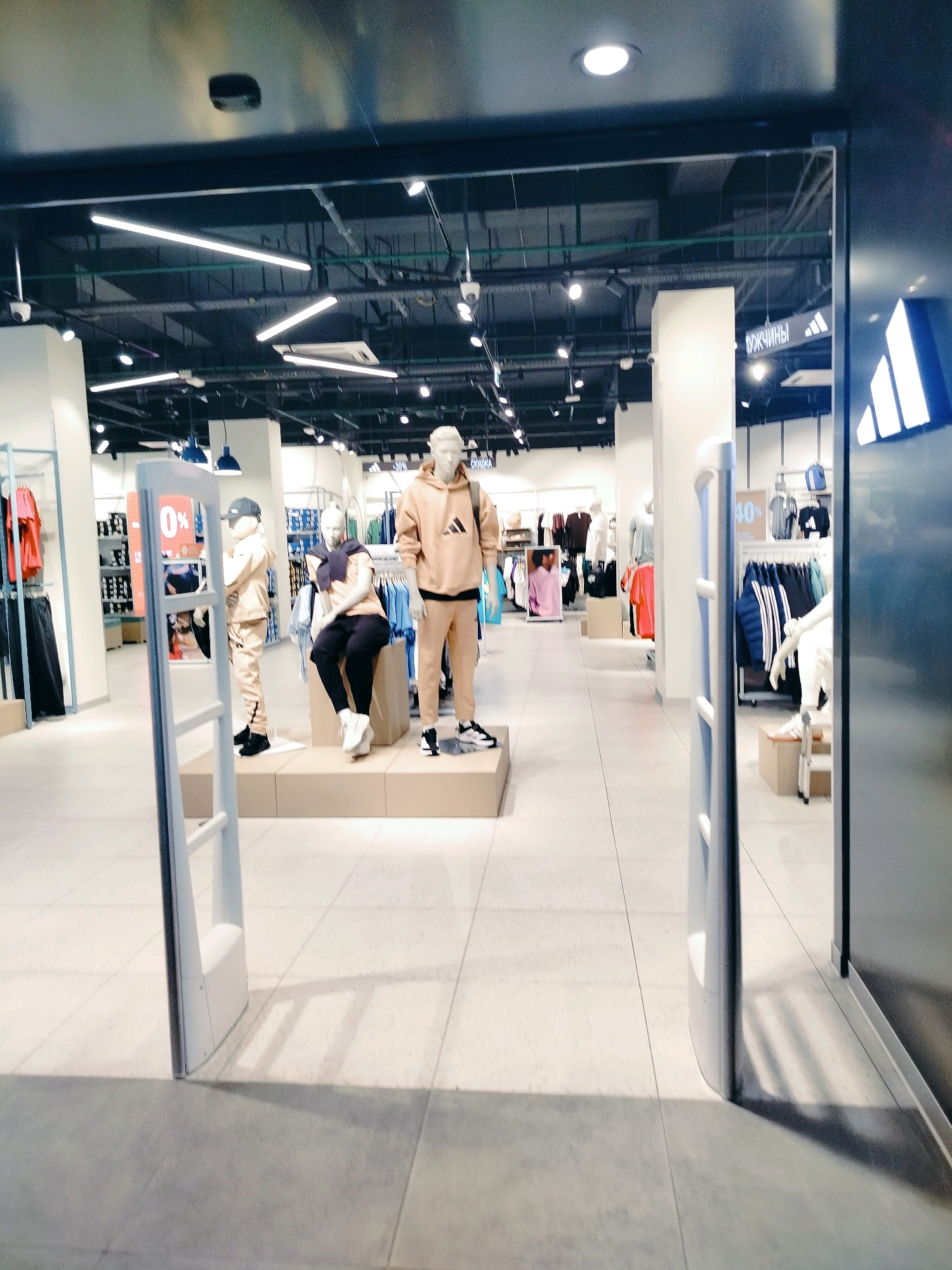 This is an indoor shot of an Adidas retail store in Shymkent, Kazakhstan. The image is taken from the entrance, looking into the store. In the foreground, there are two white security gates. Beyond the gates, the store is organized with clothing racks and displays. There are several mannequins visible, some dressed in Adidas apparel, including a male mannequin in a tan tracksuit and another in a similar outfit. Another mannequin is seated, wearing black pants and a dark blue sweater draped over its shoulders. In the background, shelves are stocked with clothing and accessories. Signs for "40%" discounts are visible on some displays. The lighting appears to be artificial, with fluorescent and track lighting fixtures on the ceiling. The floor is tiled with light-colored tiles.