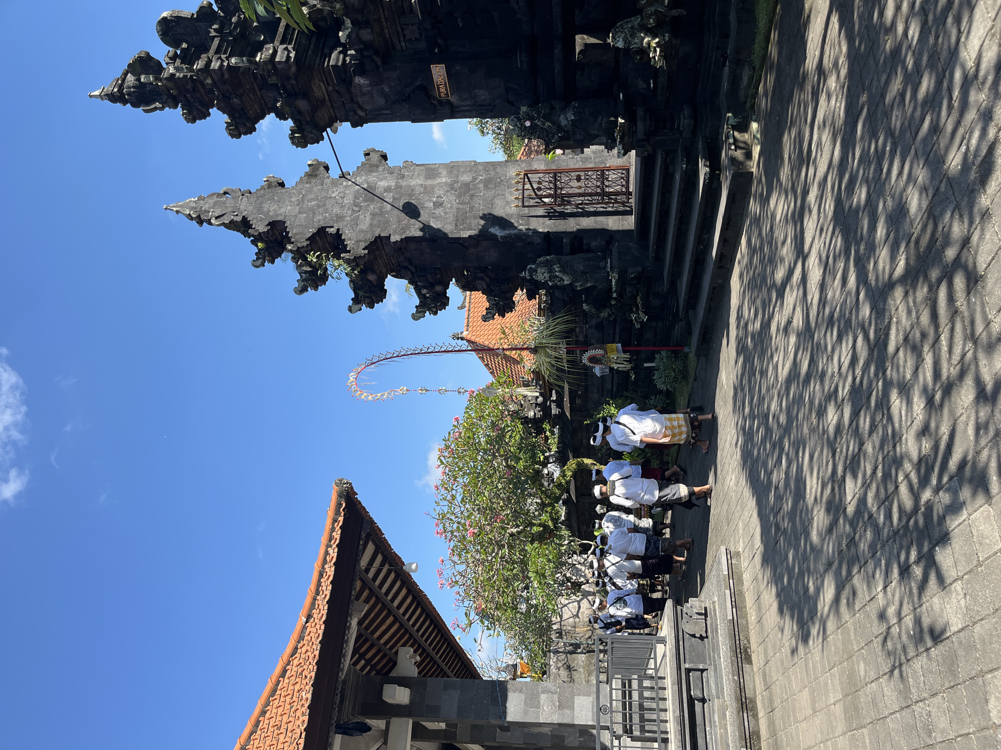 In Sukawati, Indonesia, a group of men in white shirts and sarongs walk down a paved pathway next to a temple entrance. The temple features ornate stone carvings and a tiled roof. The sky is clear and blue.