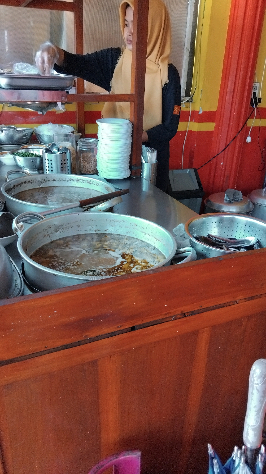 A woman wearing a yellow hijab and a black shirt is standing behind a food stall. She is wearing disposable gloves and appears to be preparing food. In front of her are several large pots of soup, a stack of white plates, and various containers of ingredients. The stall is decorated with red and yellow striped fabric. The scene appears to be a food stall in Batang, Indonesia.
