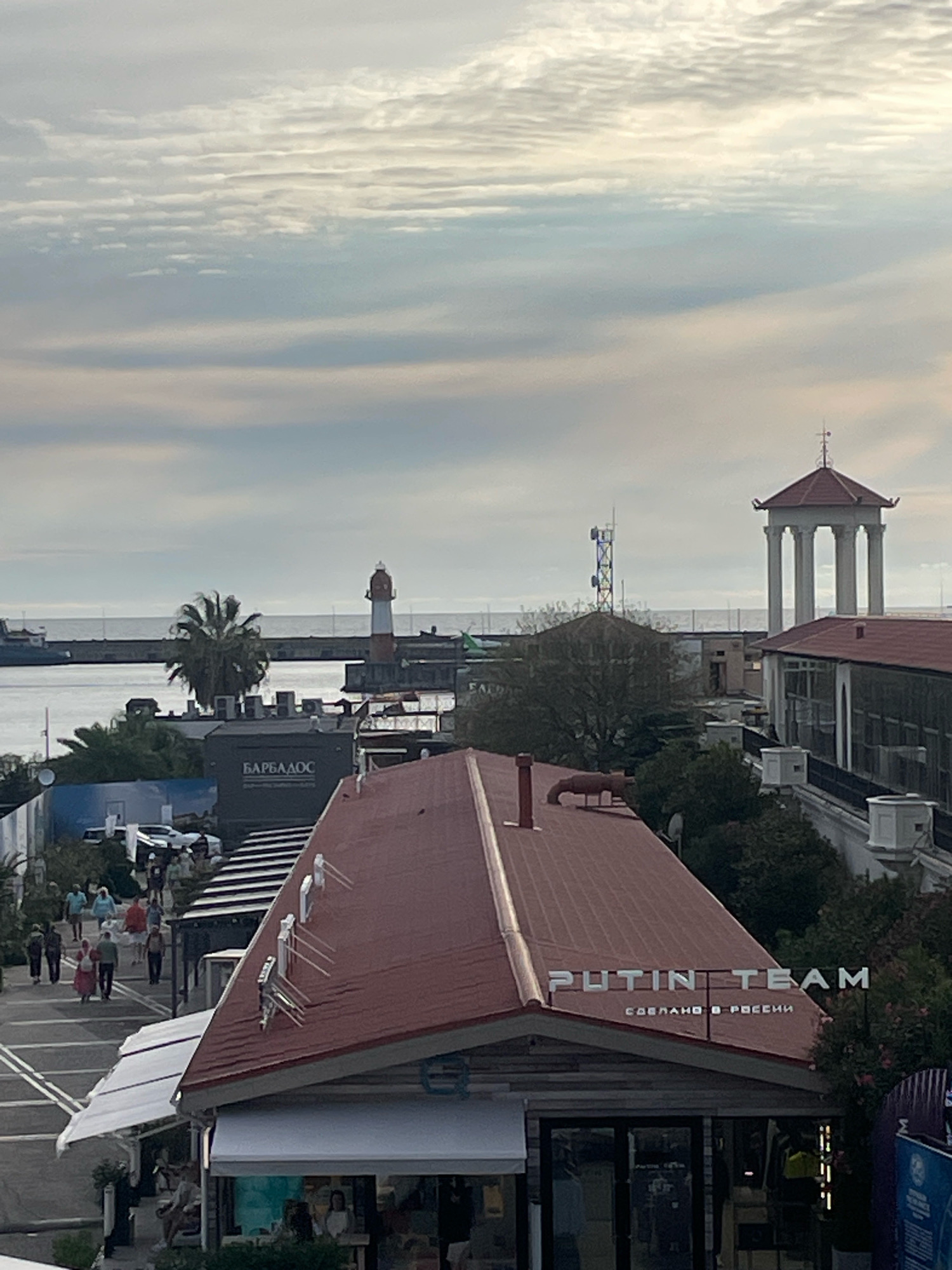 The image shows a view of a waterfront area in Sochi, Russia, likely during the day under an overcast sky. In the foreground, there is a building with a red-tiled roof. A sign on the building reads "PUTIN TEAM" in large silver letters, with smaller Russian text below that says "СДЕЛАНО В РОССИИ" (Made in Russia). There are people walking on a pathway to the left of this building, some are casually dressed.
In the middle ground, there is a building with a sign that says "БАРБАДОС" (Barbados) in white letters. A palm tree stands near this building. Further into the background, a lighthouse with red and white stripes is visible near the water. Beyond the lighthouse, there's a breakwater extending into the sea. To the right of the lighthouse, there's a structure resembling a rotunda with columns and a red roof. A telecommunications tower with antennas is also visible in the distance. The sea is calm, and the sky is filled with layered clouds, suggesting a mild or possibly cool day. The overall impression is one of a coastal city with a mix of modern and older architectural elements.