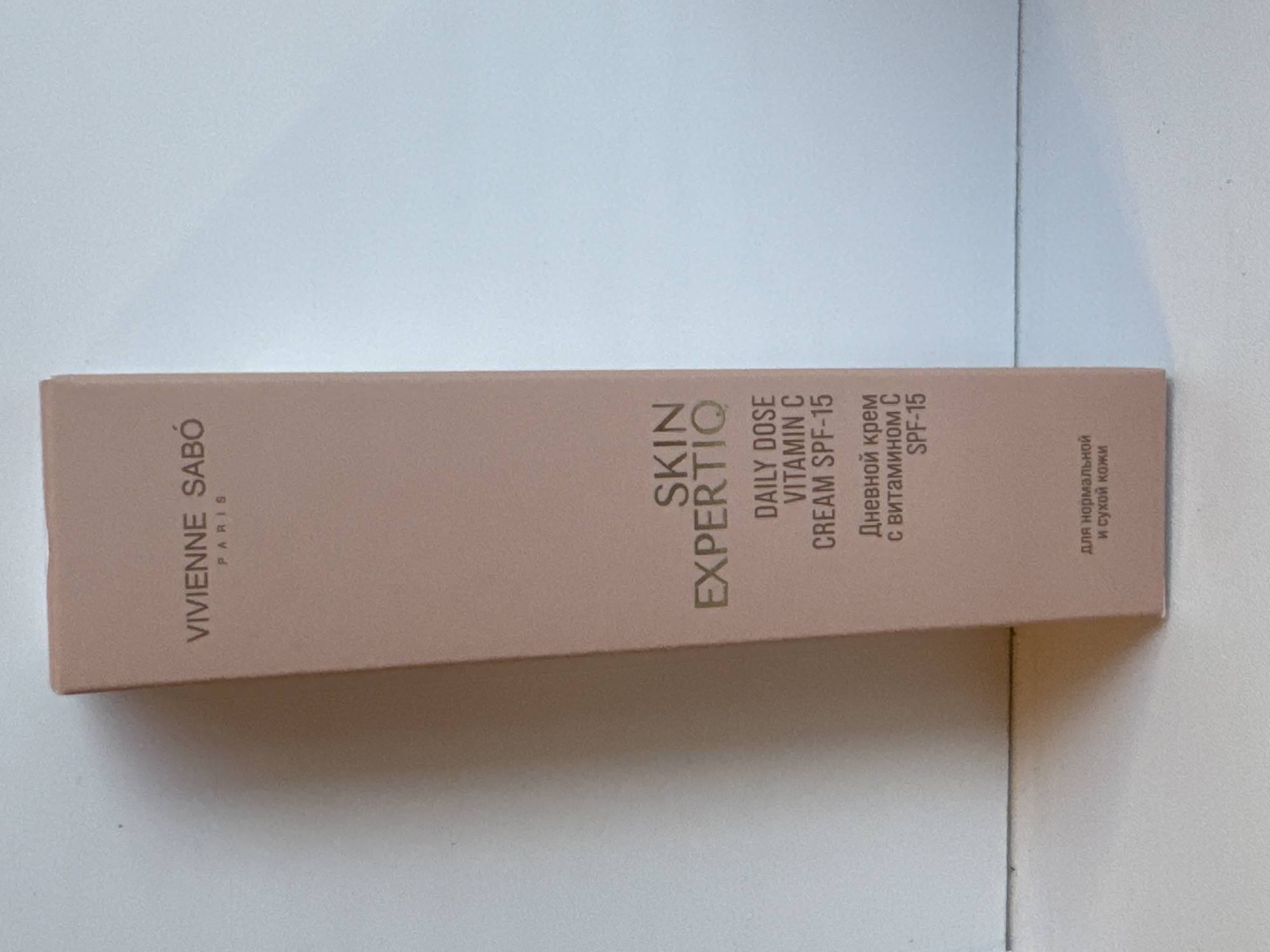 The image shows a close-up, eye-level, medium shot of a pink cardboard box for VIVIENNE SABÓ SKIN EXPERTIO DAILY DOSE VITAMIN C CREAM SPF-15. The box is oriented vertically and positioned slightly to the left of the frame's center. The background is a plain, light-colored surface, likely a wall or table, with a subtle shadow cast by the box on the bottom left. The lighting is soft and even, with no harsh shadows. The text on the box is in gold lettering. The top of the box displays "VIVIENNE SABÓ PARIS". Below that, it reads "SKIN EXPERTIO". Further down, it says "DAILY DOSE VITAMIN C CREAM SPF-15". The bottom of the box has text in Russian, which translates to "Daily cream with vitamin C SPF-15" and "for normal and dry skin". The box appears to be new and undamaged. No people or other objects are visible in the image. The scene is a product shot.