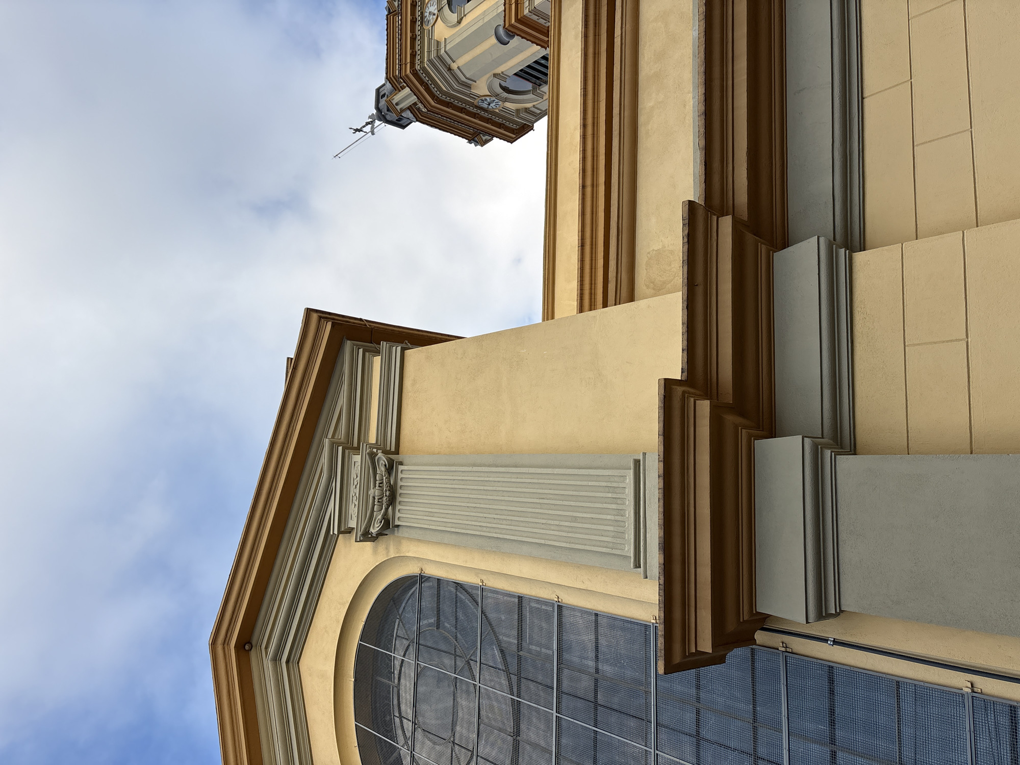 The image shows a close-up, low-angle view of the facade of a building, likely a church, with a cloudy blue sky in the background. The architecture features ornate details, including a large arched window with a metal grid, fluted columns, and layered cornices. The building's exterior is painted in warm yellow and gray tones, with brown accents on the cornices. In the upper right portion of the frame, a section of a bell tower with clocks is visible against the sky. This architectural style is typical of many churches and historical buildings in São Paulo, Brazil, particularly those with European influences. No people or specific actions are depicted in the image. The focus is entirely on the building's structure and its decorative elements.