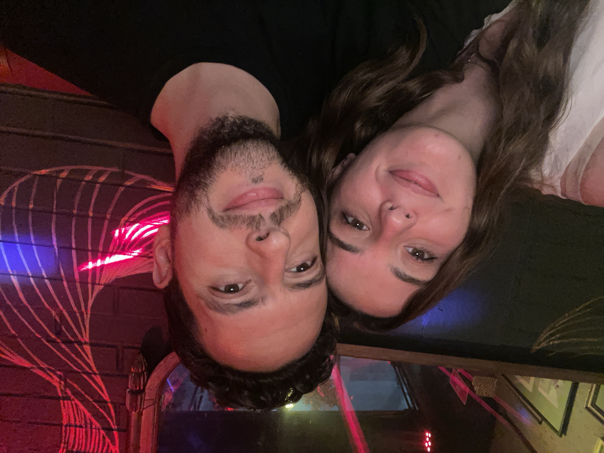 A man and woman pose closely for a photograph in a dimly lit venue, likely in Puebla, Mexico. The man has dark hair and a beard, wearing a dark top. The woman has long brown hair and wears a light-colored top. A brick wall with neon lighting is visible in the background. Other decorative elements and reflections appear in the lower portion of the frame, suggesting a bar or restaurant setting.