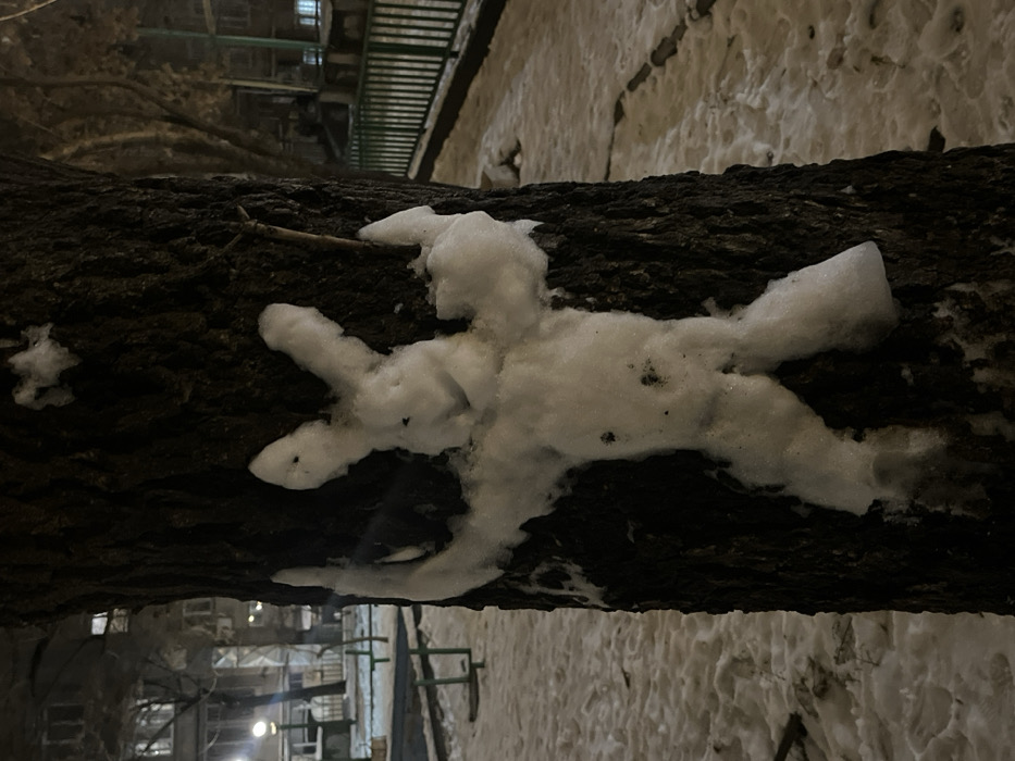 A tree trunk lies horizontally in the center. On top of the trunk is a white snow figure resembling an abstract humanoid shape with four limbs. The background is blurred and shows a snowy ground, buildings with lit windows, and metal bars, likely part of a fence or structure. The light appears to be dim, suggesting nighttime. The image is slightly rotated counterclockwise.