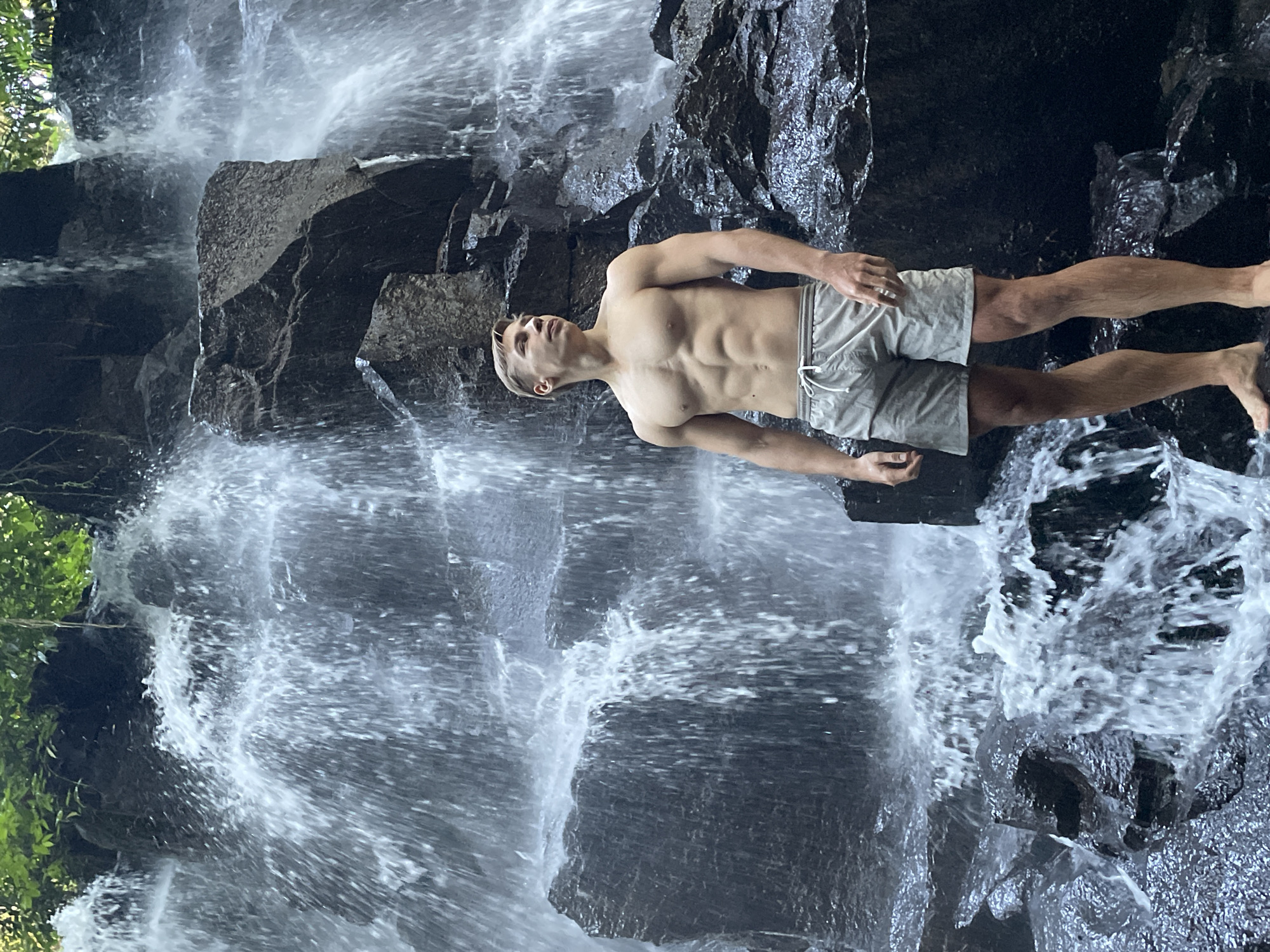 A medium shot depicts a man in his early twenties standing beneath a waterfall. The man is facing the camera with his body positioned horizontally. He is fair-skinned, has short blonde hair, and a muscular physique. He is wearing light gray shorts with a drawstring waist. Water is flowing over him and the surrounding rock face. The waterfall is situated in a dense forest, with greenery visible on the periphery. The scene is likely set outdoors. The location is near Gianyar, Indonesia.