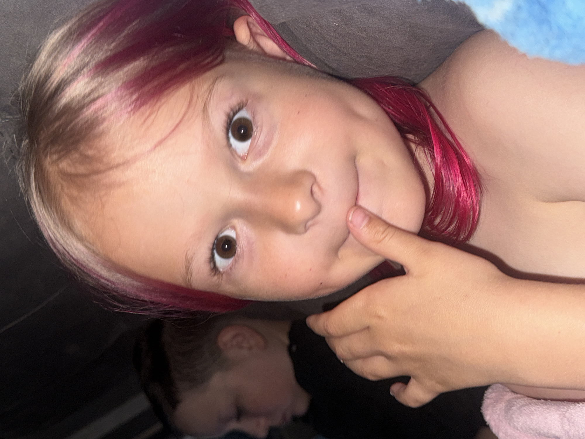 A young child with brown hair and dyed magenta streaks is lying down, looking at the camera with a playful expression. One finger is touching their cheek. A second person is partially visible in the lower portion of the frame, appearing to be lying down as well. The background consists of dark, undefined objects and surfaces. The location is unknown.