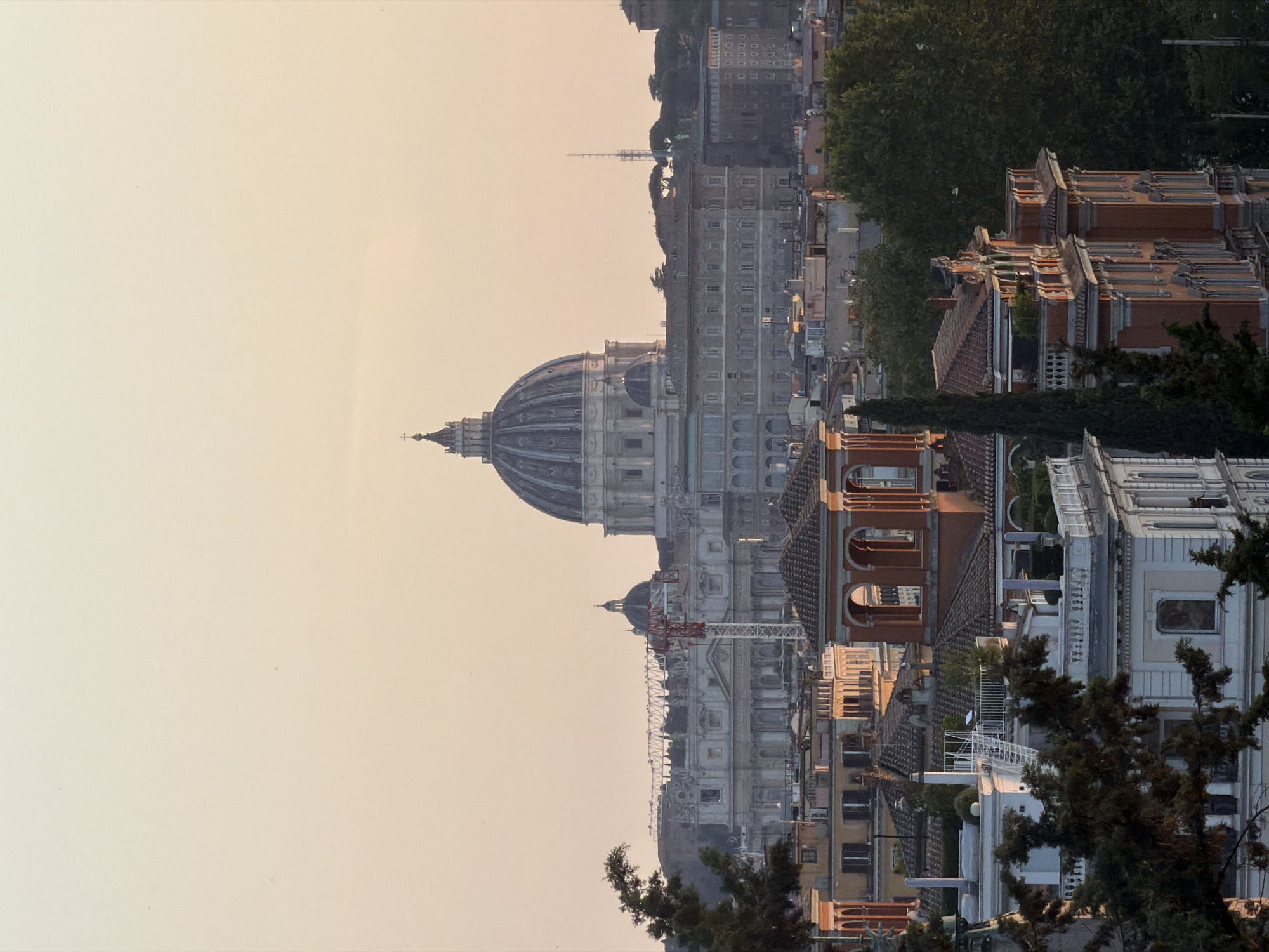 An aerial view captures the city of Rome, Italy, bathed in the soft, warm glow of sunset. The iconic dome of St. Peter's Basilica dominates the skyline, its grandeur softened by the atmospheric haze. In the foreground, a cluster of terracotta-roofed buildings with arched windows and decorative balconies are nestled amongst lush green trees. A construction crane is visible to the left of the basilica, hinting at ongoing development within the ancient city. The sky above is a gradient of pale yellow and orange, creating a serene and picturesque scene.