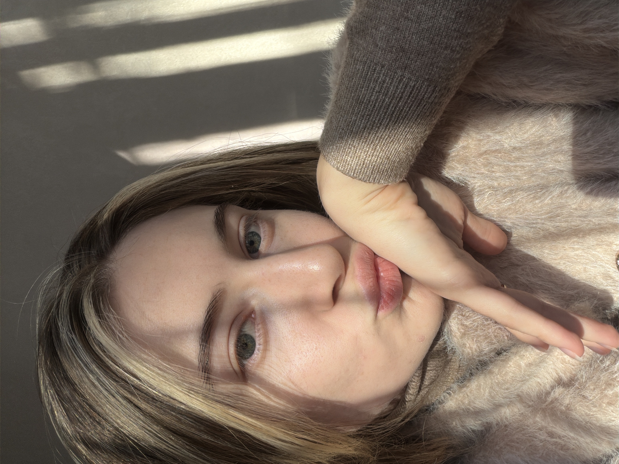 A young woman lies on her side, her face angled towards the camera. Her hair is highlighted and partially covers her left cheek. She is wearing a light brown, fuzzy sweater. Her hand is positioned near her face, fingers slightly splayed. Light from a window casts shadows on the wall behind her. There is no information available from the image that indicates the location is Plovdiv, Bulgaria.