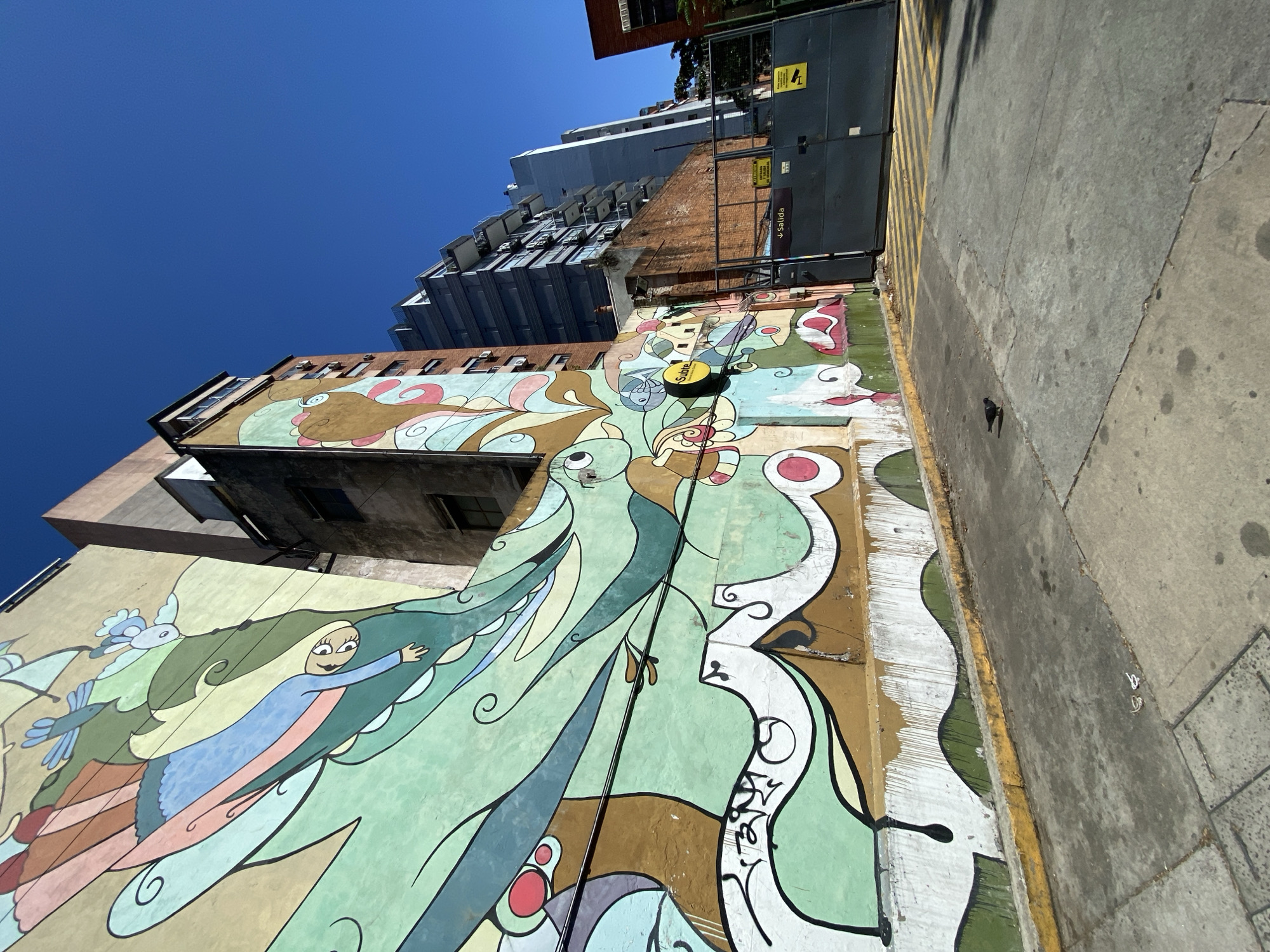 The image depicts a tall mural on the side of a building in Buenos Aires, Argentina. The mural features cartoon-like figures, including a woman with blonde hair and birds, painted in shades of green, blue, brown, and white. A modern building with glass balconies rises behind the older building with the mural. Next to the mural is a sidewalk and curb with yellow safety markings. The sky above is clear and blue.