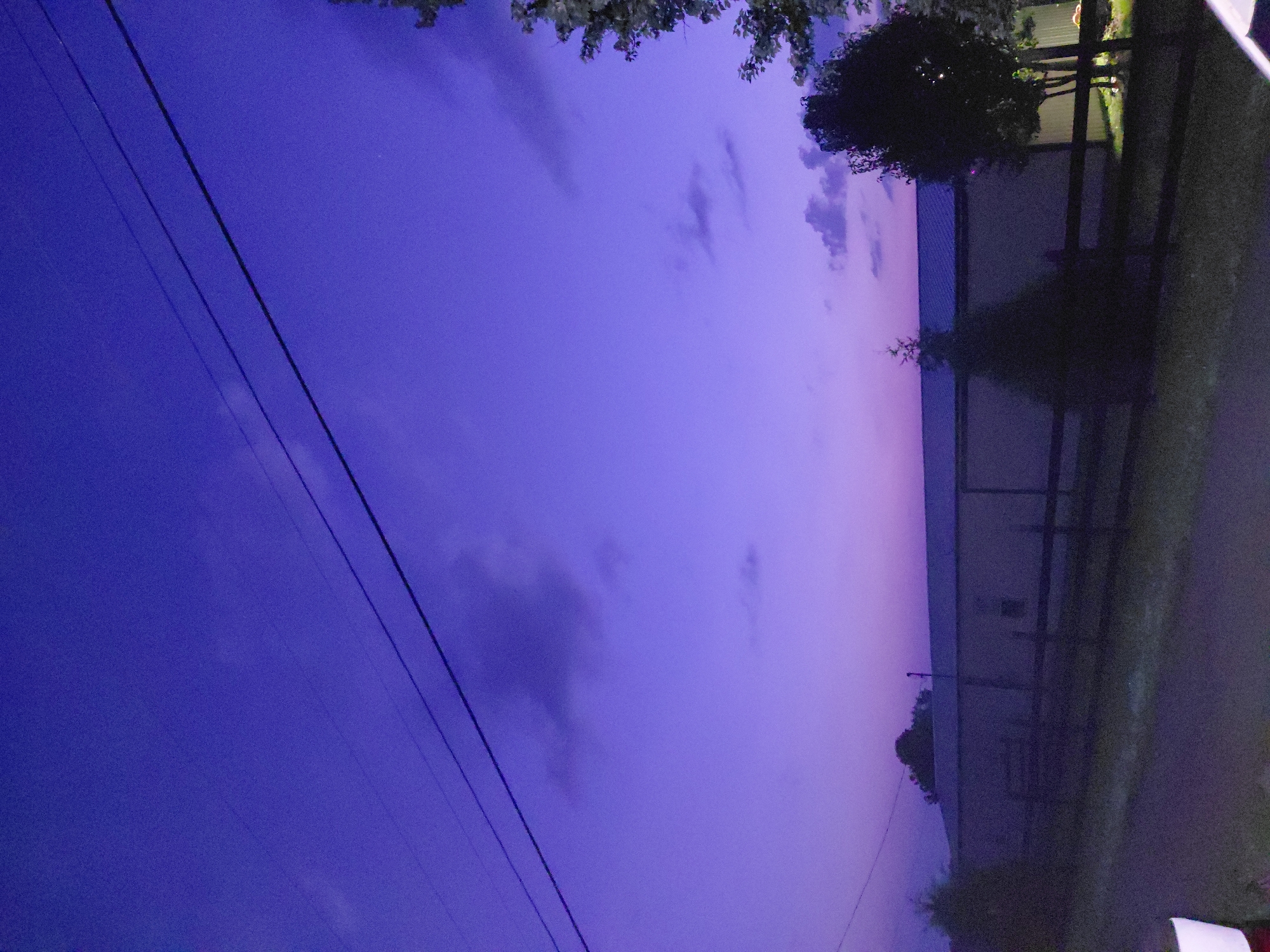 This image depicts a rural or suburban scene at dusk, characterized by a deep purple sky. Power lines are visible crisscrossing the upper portion of the frame. In the mid-ground, a long, light-colored building with a dark roof stretches horizontally. A wooden fence runs along the front of the building. Several trees, some silhouetted against the sky, are scattered across the scene, with a more prominent dark-leaved tree on the right. The foreground consists of a road or driveway. The image does not provide any specific geographical markers to indicate its location within Richmond, United States. There are no people or discernible actions occurring in the scene.