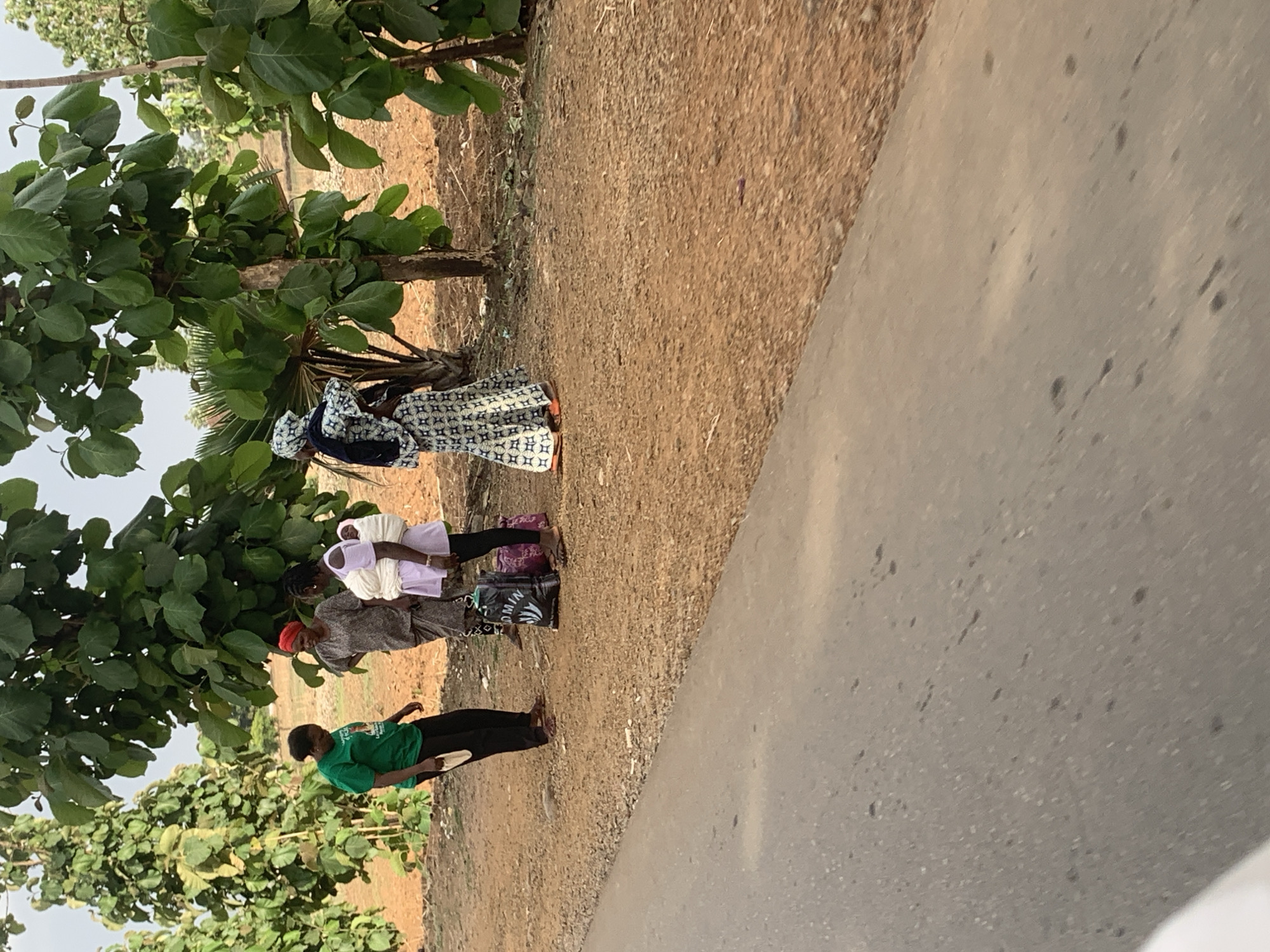 The image shows four individuals standing near a roadside in what appears to be a rural area in Maigizo, Nigeria. The location is adjacent to a paved road with sandy or dirt shoulder. Lush green foliage and trees are visible.
The individuals are standing in a vertical line, parallel to the edge of the road. From furthest away, they appear to be:
1. An adult woman wearing a long, patterned dress and head covering
2. A child in a white garment
3. An adult woman in a gray top and red head covering
4. A young person wearing a green shirt and black pants
It is not possible to determine their actions or intentions from this single image.