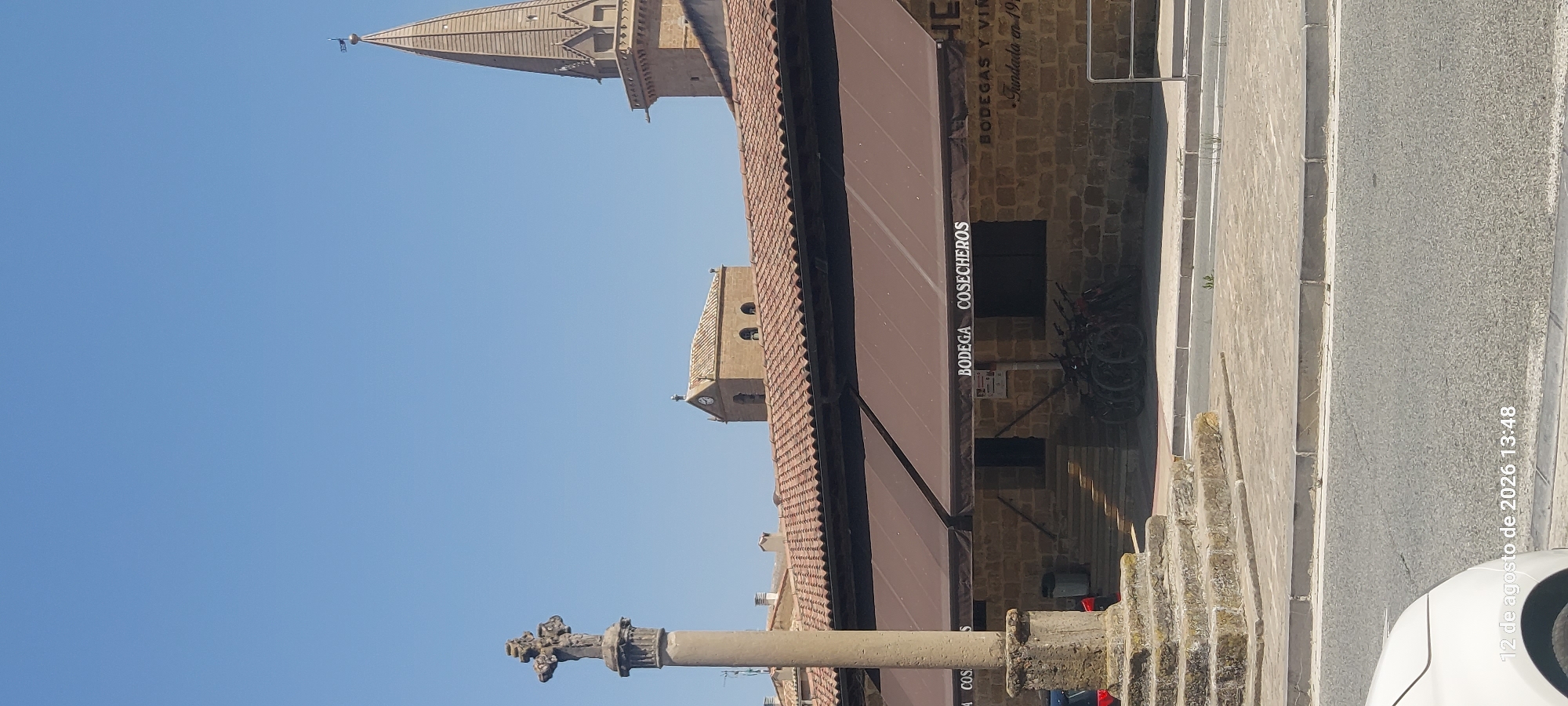 This is a vertical, eye-level, full-shot photograph taken on a clear, sunny day in Olite / Erriberri, Spain. The scene depicts a street view featuring a stone monument on the left, a building with a brown awning, and two church towers in the background. The stone monument, possibly a cross, is situated on a multi-tiered stone base. The building, constructed of light-colored stone blocks, has a brown awning stretched across its facade, displaying the text "BODEGA COSECHEROS" and "BODEGAS Y VIN". Several bicycles are parked in front of the building. In the background, two distinct church towers are visible against a clear blue sky. The tower on the left is shorter and rectangular with a tiled roof and a clock face, while the tower on the right is taller, conical, and more ornate, topped with a weather vane. The ground in front of the building is paved with light-colored stone slabs. A portion of a white car is visible in the lower left corner, partially obscuring a timestamp that reads "12 de agosto de 2026 13:48". The overall environment suggests an urban or town setting.