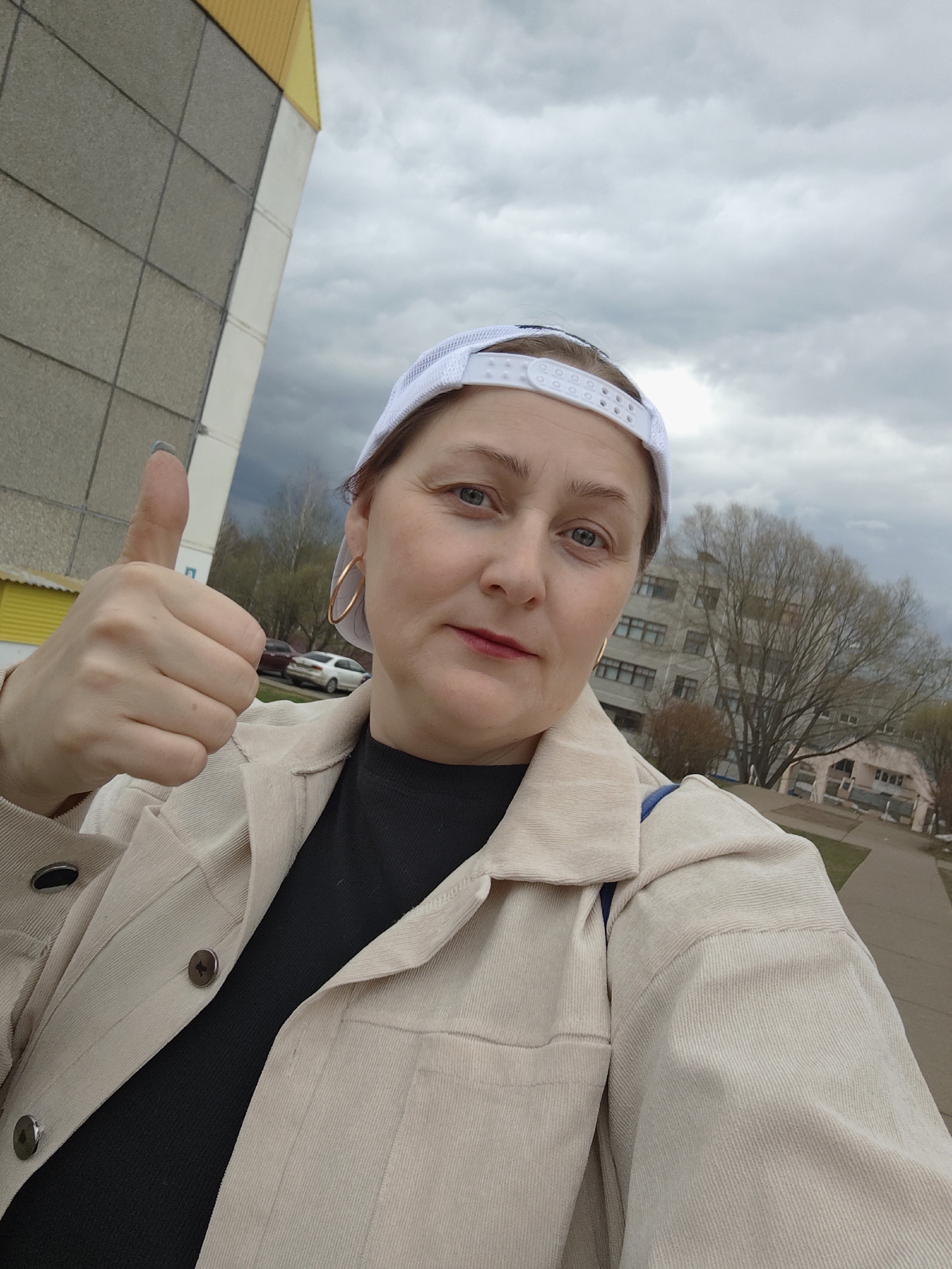 A woman with fair skin and light eyes is centered in the image, giving a thumbs-up. She wears a white baseball cap, gold hoop earrings, a black top, and a beige corduroy jacket. Behind her is a cloudy sky and a building with grey and yellow panels on the left side. Further back is a multi-story building and bare trees. The location is Марковский, Russia.