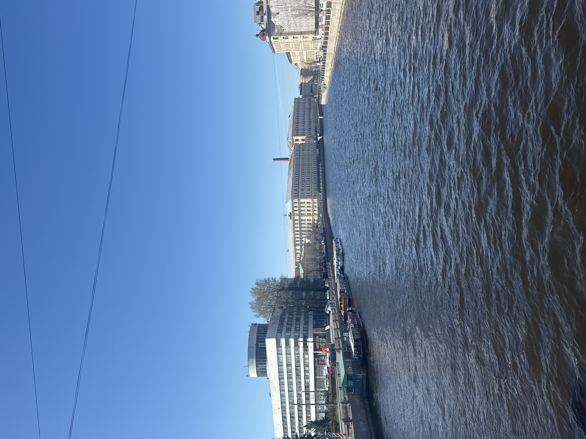 This image depicts a clear, sunny day along a wide river or canal in a city, likely Saint Petersburg, Russia, viewed from an elevated perspective such as a bridge. The left half of the frame is dominated by a bright, cloudless blue sky, traversed diagonally by two thin power lines or overhead cables.
The right half of the image showcases the city's waterfront. A broad, dark body of water with visible ripples occupies the lower portion, suggesting a moving current or wind. Several boats, possibly tour vessels or barges, are docked along the riverbank.
The bank is lined with various buildings. In the foreground, a prominent modern building with a curved facade and numerous windows stands out in white. Next to it, several trees with bare branches indicate late winter or early spring. Further along the embankment, a long, multi-story building with a tall, reddish-brown industrial chimney is visible. In the distance, a series of older, more classical-style buildings with light-colored facades line the waterfront, some showing signs of renovation with scaffolding.
There are no people clearly visible. The time of day is bright daylight, characterized by the clear blue sky and strong sunlight. Notable details include a blue parking sign with a white 'P' visible near the modern building, and what appear to be Russian national colors (red, white, and blue) on a small structure or flag near the docked boats. The overall scene captures a tranquil, observational view of a historic urban landscape.