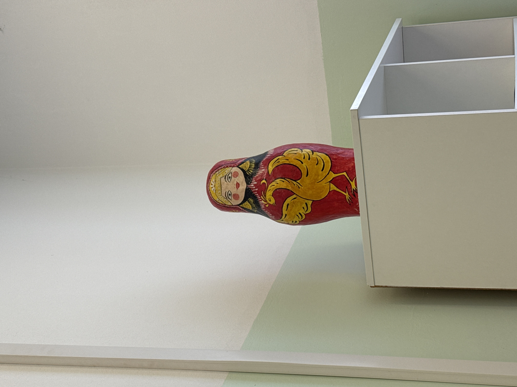 A red, matryoshka doll with yellow markings lies sideways on a two-toned wall adjacent to a rectangular, off-white shelving unit. The lower half of the wall is a light sage color, and the upper half is off-white. The frame of the shelving unit is visible, but it appears to be empty. The setting or location is unknown.
