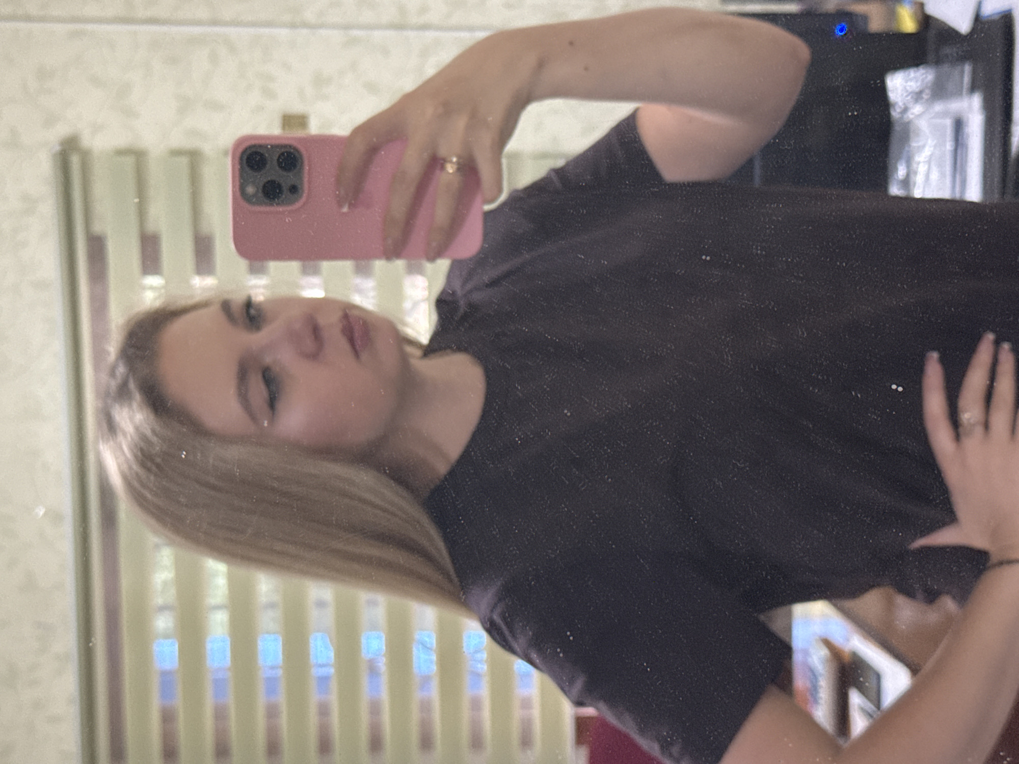 In a reflective surface, a young woman with blonde hair is captured from the waist up, holding a pink smartphone in her right hand to take a mirror selfie. She is wearing a dark, possibly black, short-sleeved t-shirt with a subtle speckled texture. Her left hand is placed on her hip. The background, also reflected in the mirror, shows a window with horizontal blinds, revealing a soft, diffused light. The overall setting appears to be indoors, possibly a domestic or personal space, given the nature of the selfie and the visible background elements. The woman's facial expression is neutral, and she is looking directly at the camera.