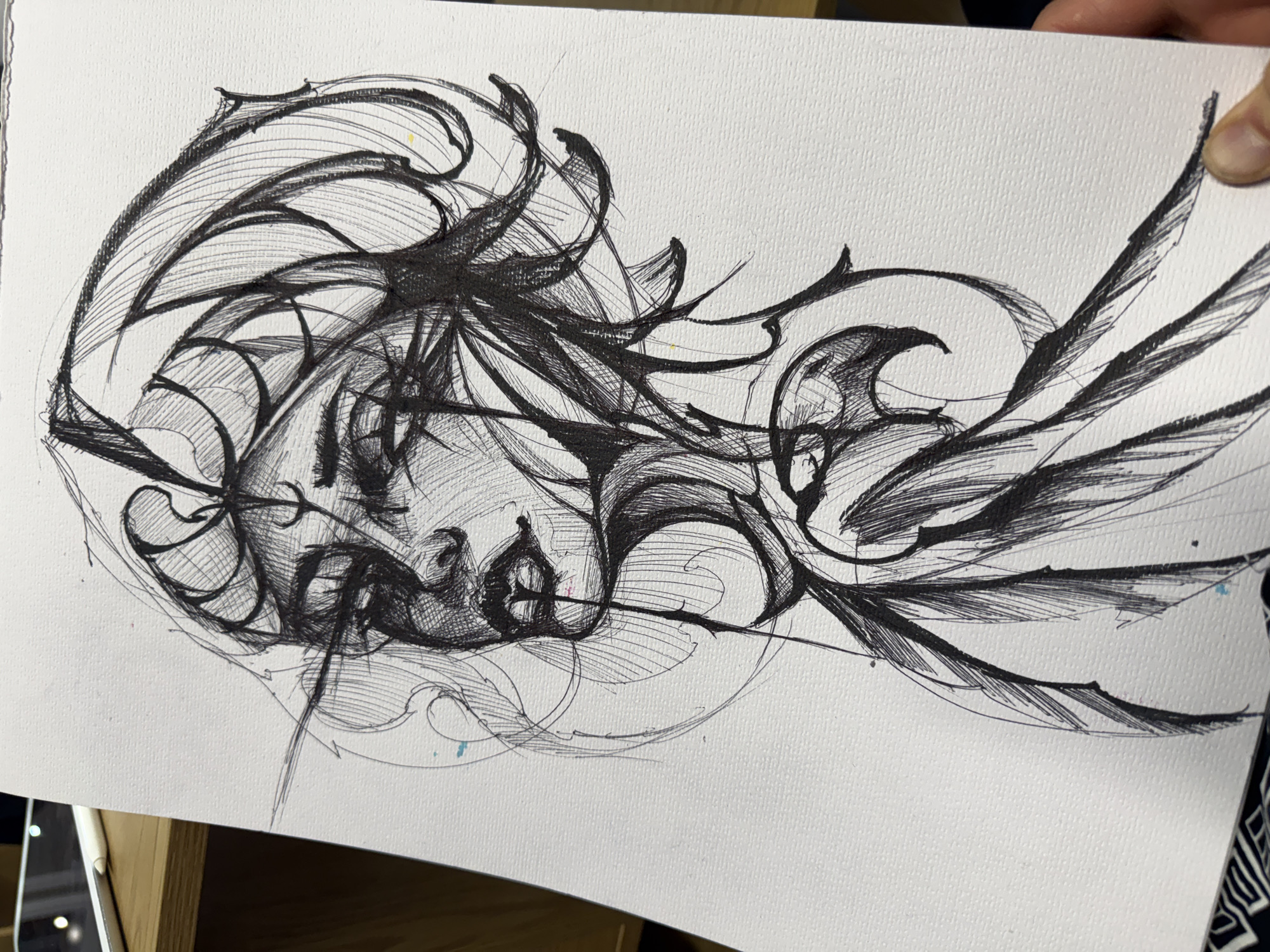 The image shows a black and white ink drawing on white paper. The drawing depicts a stylized face of a person, likely female, in a somewhat abstract and surreal style. The face is tilted to the side, with closed eyes and detailed shading creating the impression of depth and texture. Elaborate, swirling lines and shapes surround the face, acting as both a decorative element and possibly representing hair, wind, or other abstract concepts. The artwork is held in front of what appears to be a wooden surface, suggesting it is a work in progress or recently completed. There are no indications of the location being in Sofia, Bulgaria.