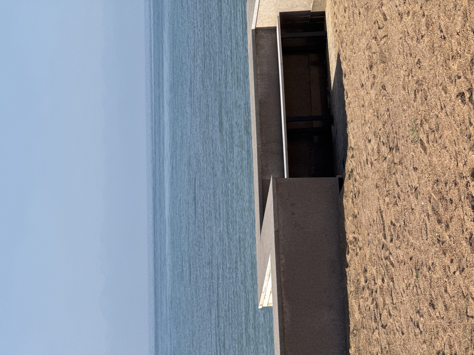 A wide shot captures a serene beach scene with a stone embankment and the Issyk-Kul lake under a hazy sky. The sand in the foreground is a light brown, scattered with small pebbles and imprints of what appear to be animal tracks. To the left, a light brown, angular stone structure forms a low wall, with another similar wall extending to the right, creating a recessed area. Within this recessed area, a long, narrow bench with a light-colored top and dark metal legs is visible, suggesting a public seating area. Beyond the embankment, the tranquil, blue waters of Issyk-Kul stretch out, meeting the pale blue, hazy sky at the horizon. In the far distance, faint mountain silhouettes are barely discernible through the atmospheric haze, indicating the mountainous terrain surrounding the lake. The lighting suggests it is daytime, possibly late morning or early afternoon, with the sun casting subtle shadows. The overall atmosphere is peaceful and quiet, characteristic of a beach resort area.