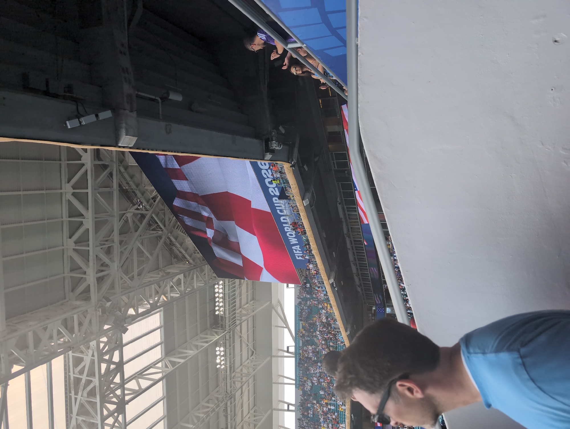An outdoor stadium scene in Miami Gardens, Florida, United States of America, is depicted from a low-angle perspective. The foreground shows the back of a man wearing sunglasses and a light blue t-shirt, looking upwards. Beyond him, a large stadium screen displays a close-up of an American flag, with the text "FIFA WORLD CUP 2026" prominently visible. The screen is mounted on the underside of a stadium structure, with white support beams visible above. In the mid-ground, tiered seating filled with spectators can be seen, some individuals are interacting with each other. A man and a woman are standing on a staircase in the mid-ground, facing each other, with the man appearing to gesture. The overall environment suggests an event or game is taking place within the stadium.