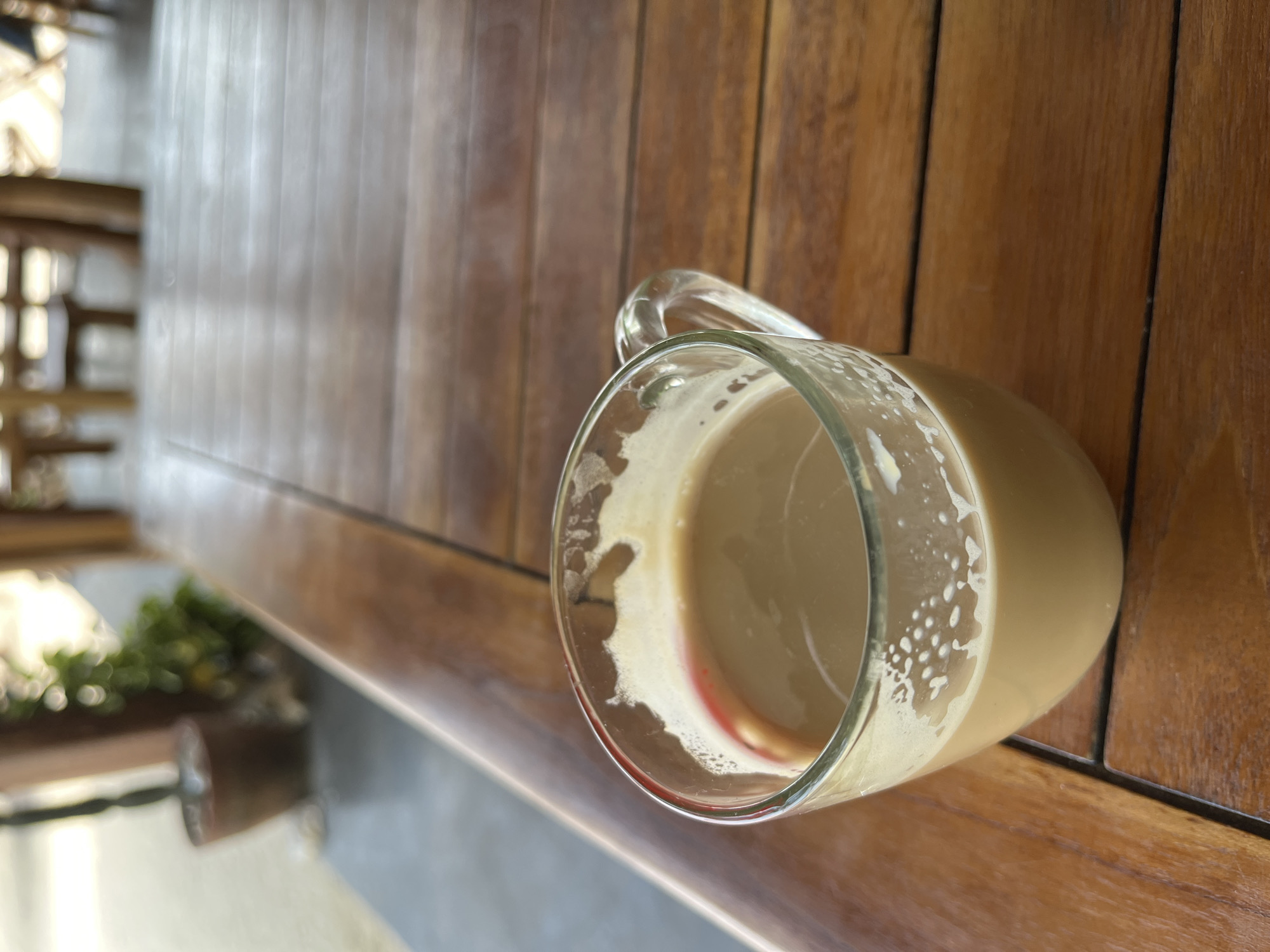 A close-up shot focuses on a clear glass mug filled with a light brown liquid, likely coffee with milk, placed on a wooden table. The mug has a handle and a frothy layer on top, with some dried residue visible on the rim, including a faint red mark on the upper edge of the glass. The table's surface is made of polished wooden planks with visible grain. In the background, the scene is out of focus, hinting at an outdoor or semi-outdoor setting with wooden furniture and greenery. The overall atmosphere suggests a relaxed moment, possibly at a cafe or a restaurant in Legonipah, Indonesia.