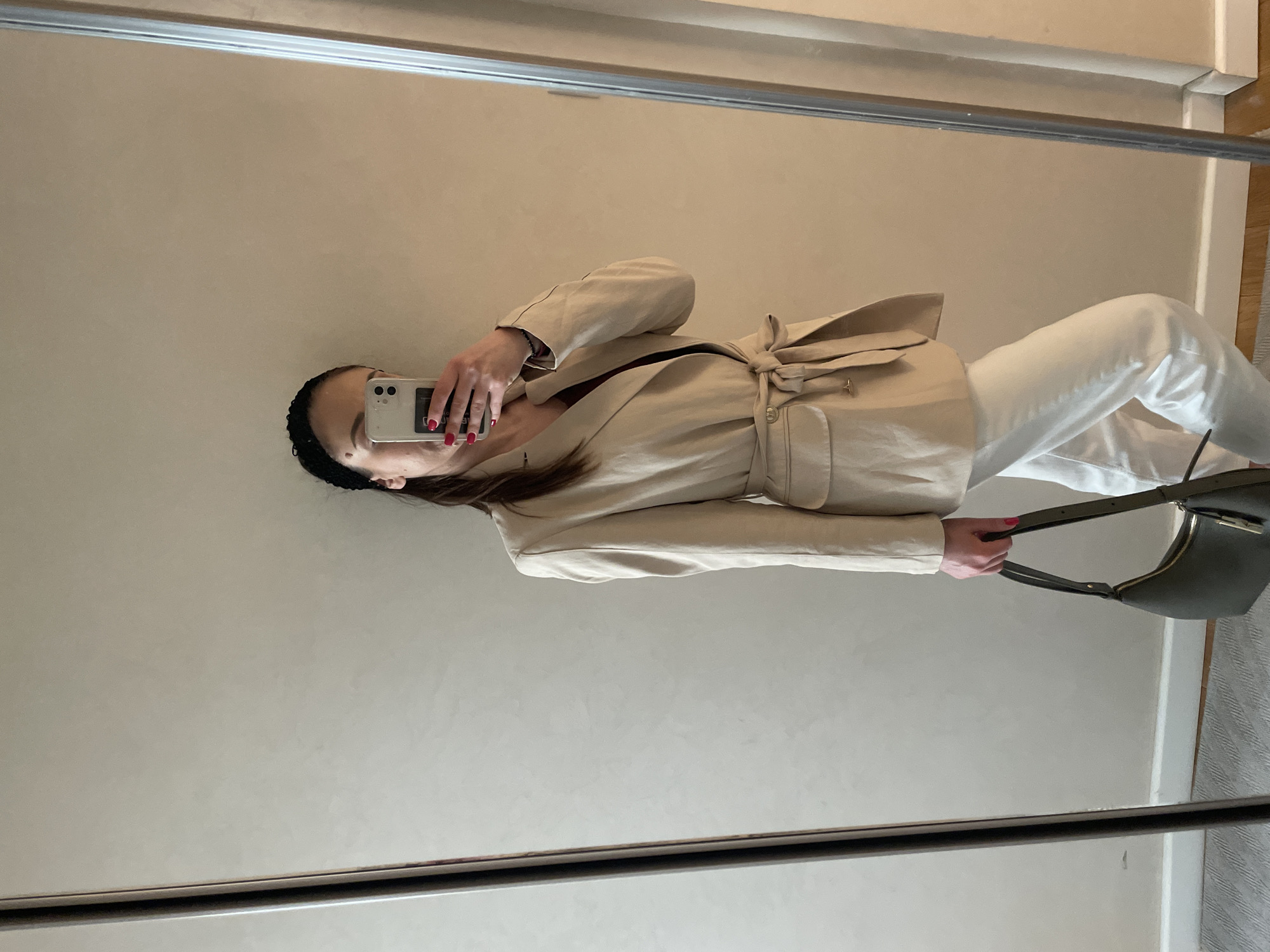 A person in a beige jacket and white pants is taking a selfie in a mirror. They are holding a smartphone in their left hand and a dark green handbag in their right. The mirror is framed and reflects a portion of the wall and what seems to be part of the room's interior, including a glimpse of wooden trim. The background appears to be a neutral-toned wall. The location of this photo in the city of Varna, Bulgaria is not possible from this image.