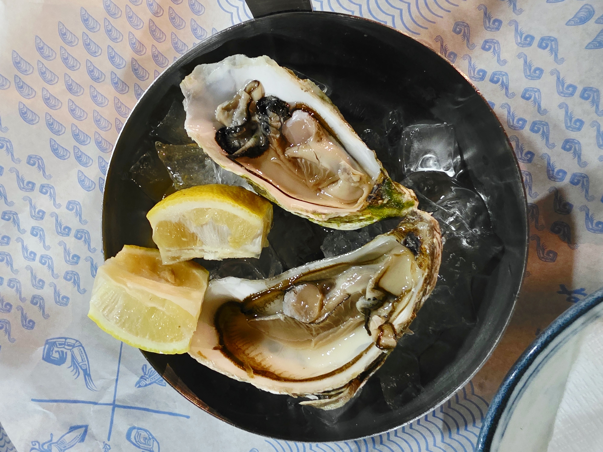 Two oysters are served on ice in a black circular dish, garnished with three lemon wedges. The dish is placed on a white paper liner with a blue decorative pattern of stylized sea creatures and mussels. The scene appears to be an indoor setting, possibly a restaurant, given the presentation of the food. The lighting suggests it is daytime or under bright artificial lights. There are no people visible in the image. The specific location in Larnaca, Cyprus, cannot be determined from the visual information provided.