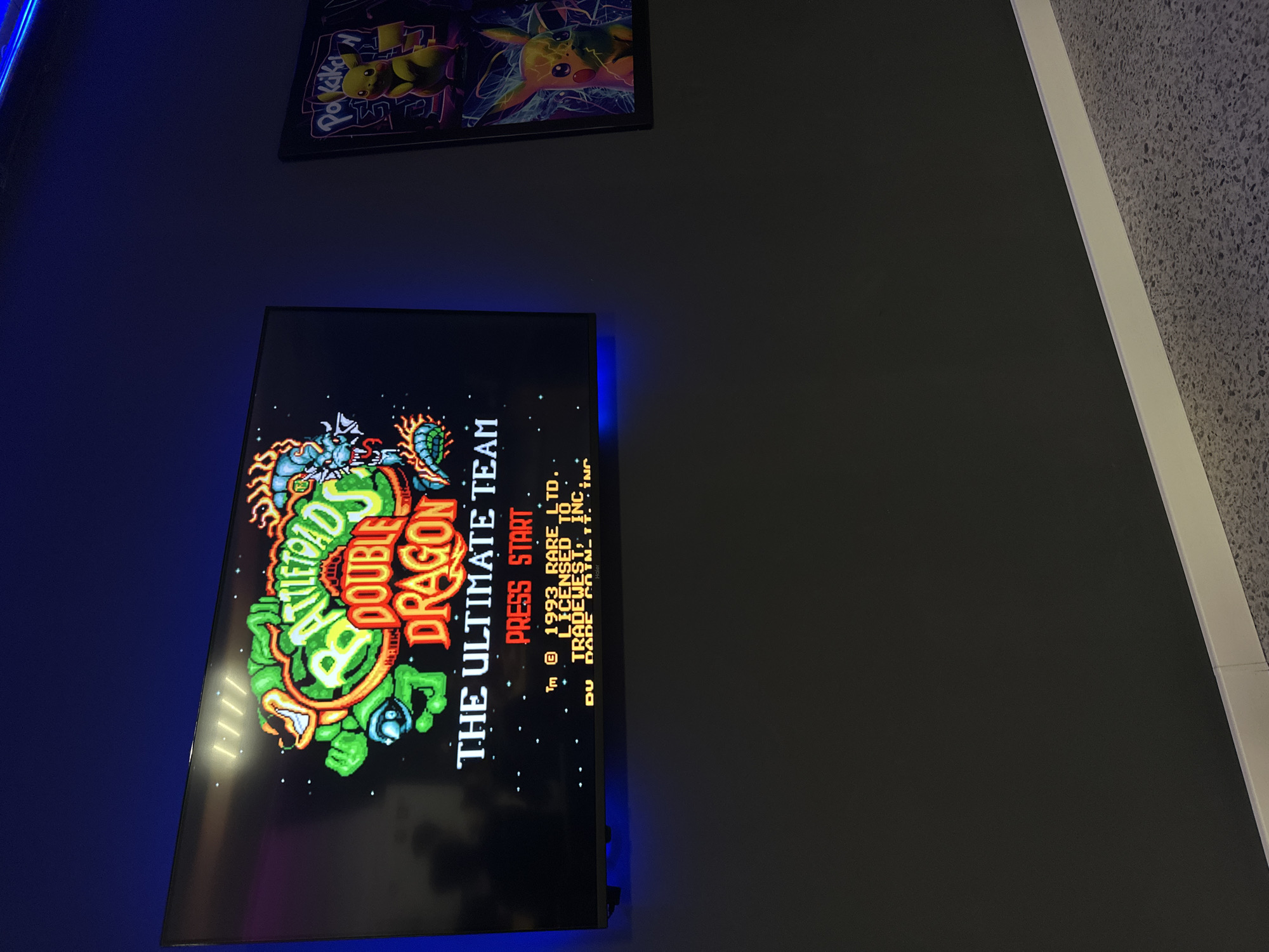 The image shows a television screen displaying the title screen of the video game "Battletoads Double Dragon: The Ultimate Team". The screen is mounted on a dark wall. Above the television is a framed picture, possibly featuring Pokémon characters. The wall to the right of the television is a lighter grey or white. The scene is indoors.