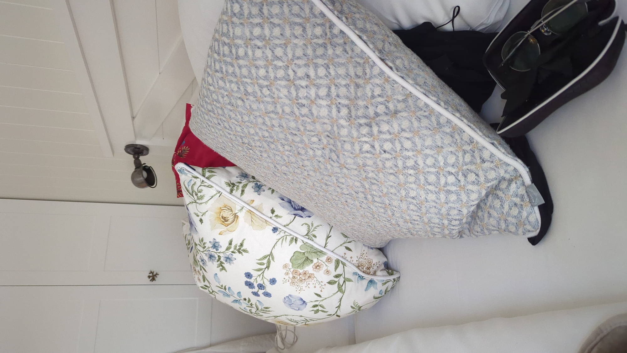 The image depicts a close-up view of a bed in what appears to be a bedroom. Two decorative pillows are prominently featured. The pillow in the foreground, to the right, is rectangular and covered in a light blue and white woven fabric with a subtle geometric pattern. It has a white piping detail along its edges. The pillow behind it, to the left, is also decorative and has a floral pattern with blue, yellow, and green elements on a white background, also with white piping. Between these two pillows, a portion of a red pillow is visible.
The bed appears to be made with white linens. Behind the pillows, a white headboard with horizontal panels is visible. To the left of the bed, a portion of a white wardrobe or closet door is visible, with a decorative handle or embellishment. Above the pillows and headboard, a small, dark metal wall-mounted light fixture with an adjustable arm is attached to a cream-colored wall with vertical wooden planks.
In the lower right corner of the image, a dark-colored eyeglasses case is open, revealing a pair of round-lensed glasses with thin metal frames. A piece of black fabric, possibly clothing, is tucked beneath the blue and white pillow.
The lighting suggests an indoor setting, likely during the daytime, with natural light possibly supplemented by the wall fixture. The overall impression is one of a neatly arranged, possibly domestic space. The location of Larkollen, Norway, is mentioned as context for the scene, implying this is a bedroom within a residence in that area.