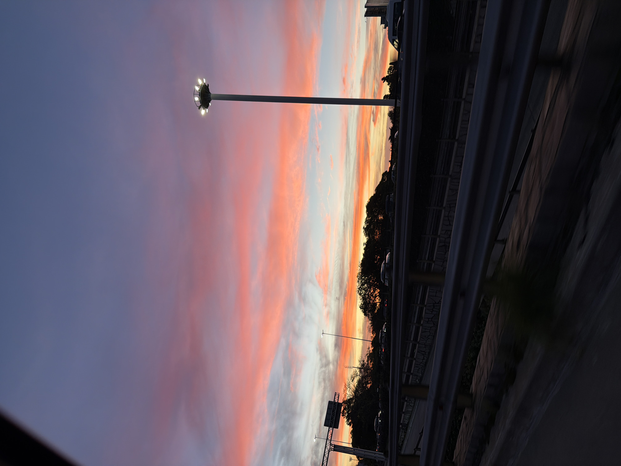 The image shows a sunset sky in Buenos Aires, Argentina, featuring hues of purple, pink, and orange. A tall light pole with multiple bulbs is visible on the left. A highway with metal guardrails runs horizontally across the lower half of the image, with trees and vehicles visible beyond the guardrails. Road signs are also present. The photograph appears to have been taken from a vehicle.