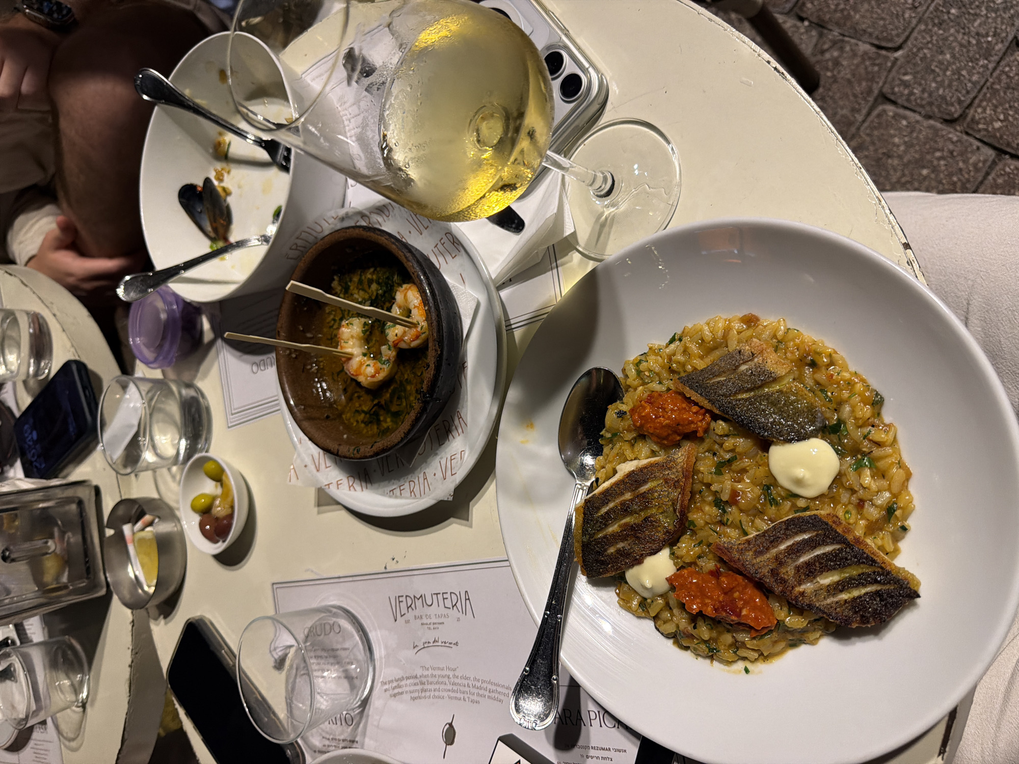 The image depicts a table at a restaurant in Tel-Aviv, Israel, likely at "El Taller de tapas" based on the menu visible in the foreground. The scene captures a meal in progress, with several dishes and drinks on the table. In the foreground, a white plate holds a portion of risotto topped with pieces of grilled fish, dollops of mayonnaise, and red chili paste. Adjacent to this is a small, dark bowl containing two shrimp on skewers swimming in oil and herbs, and a larger white bowl with remnants of mussels and forks. A glass of white wine, partially filled, sits to the right of the bowl of shrimp, with a smartphone visible underneath its base. Other items on the table include a small bowl of olives and lemon slices, glasses of water, a silver grater, and a menu. The lighting suggests an evening setting, possibly outdoors, with a cobblestone surface visible beneath the table. The overall impression is one of a casual dining experience.