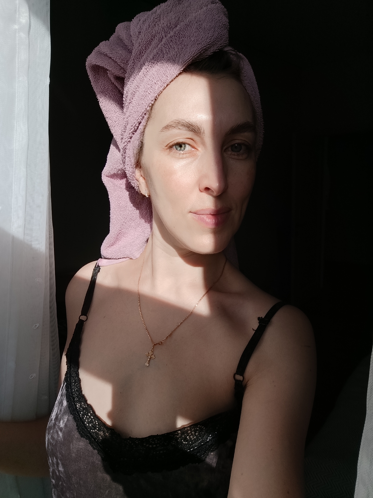 A young woman stands indoors near a window. She has a towel wrapped around her head, and is wearing a camisole top. A gold necklace with a cross pendant is visible. The left side of her face is illuminated by sunlight, while the right side is in shadow. The background consists of a sheer white curtain on the left and a dark area on the right. The picture was taken in Chaikovsky, Russia.