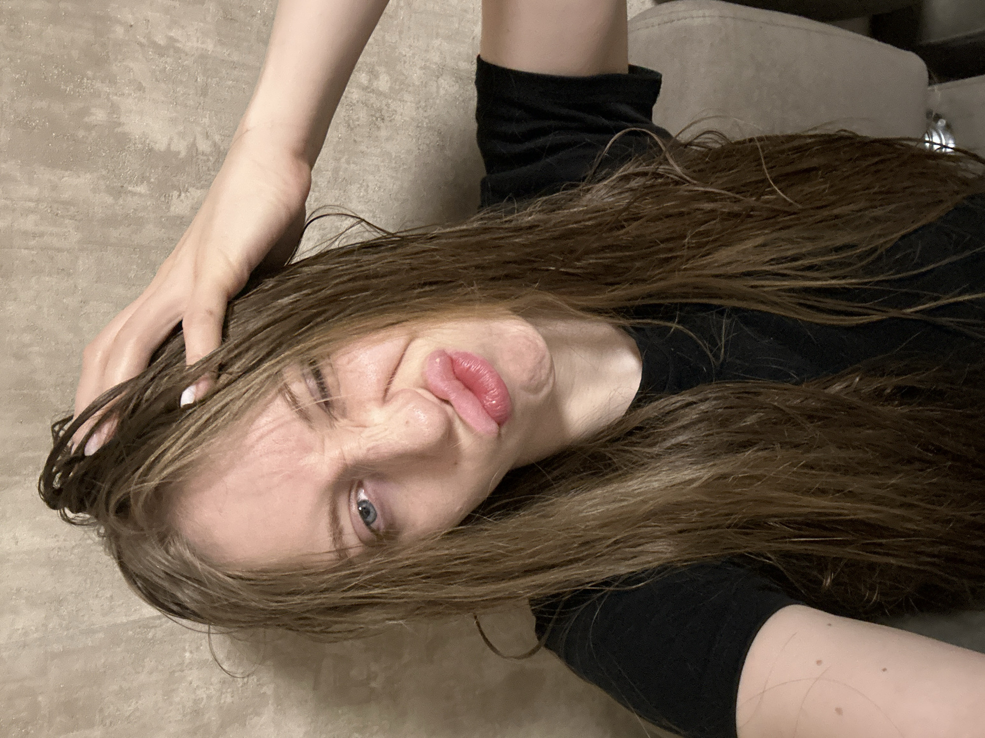 A woman with long, wet brown hair is lying on a beige surface, possibly a sofa or floor, indoors. She is wearing a black t-shirt. Her head is tilted, and she is making a pouting face with her lips. One hand is touching her hair. The background is blurred. It is not possible to confirm the location as Chaikovsky, Russia with the information available.