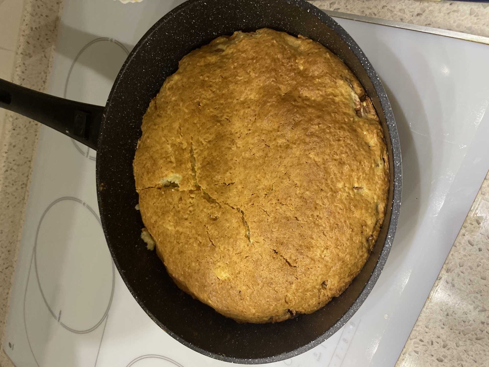 A top-down, medium shot depicts a cooked round cake in a black non-stick frying pan on top of a white ceramic cooktop. The cake's surface is golden brown with visible cracks. The handle of the pan extends to the left. The cooktop is situated on a countertop with a speckled light brown and white pattern. The image was taken in an indoor environment, most likely a kitchen, in the city of Moskovskiy, Russia.