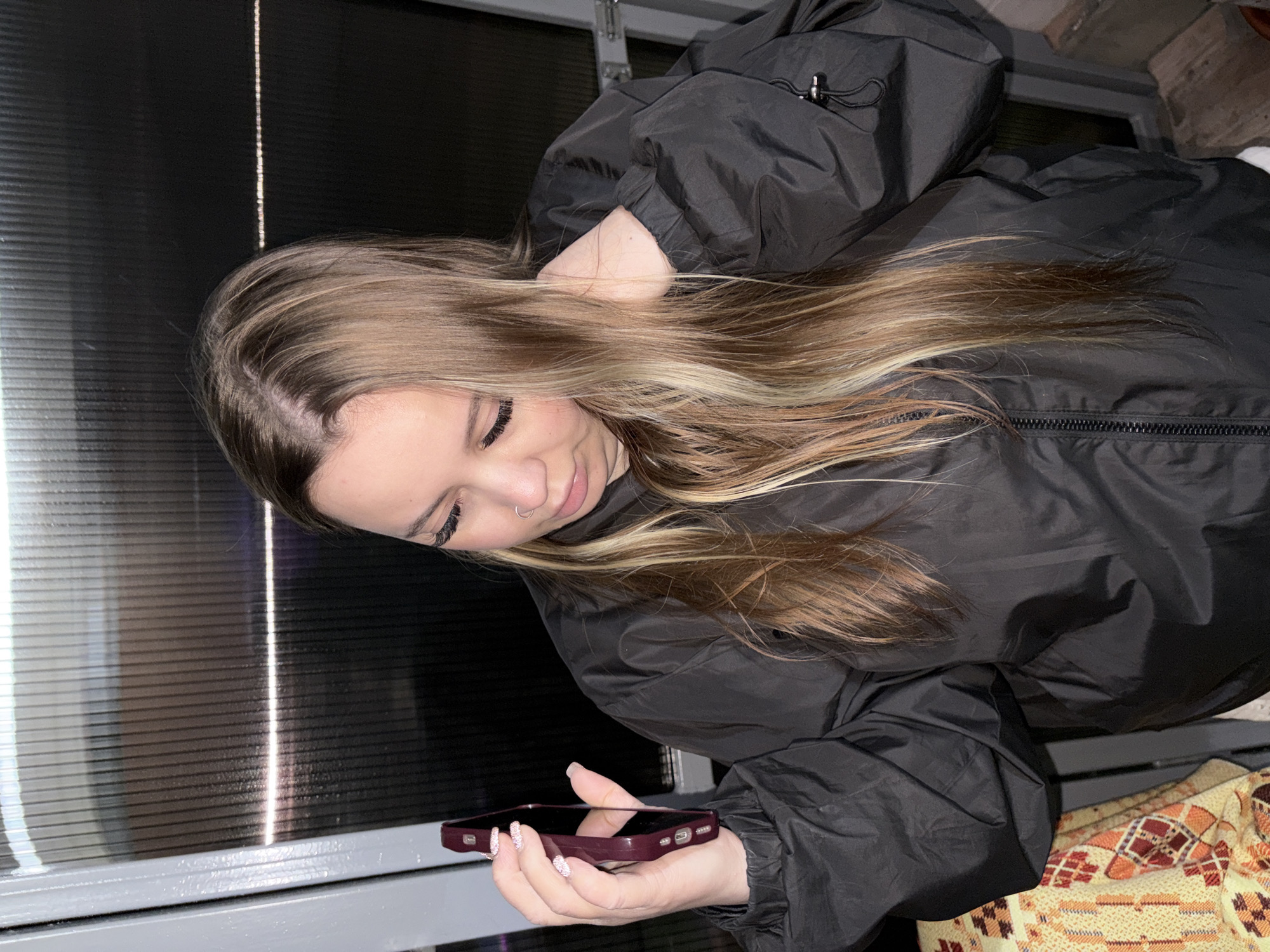 A young woman with long blonde hair is holding a dark-colored smartphone. She is wearing a black puffy jacket. The background includes a window or wall with horizontal lines and a patterned fabric. The location is Novopokrovskaya, Russia.