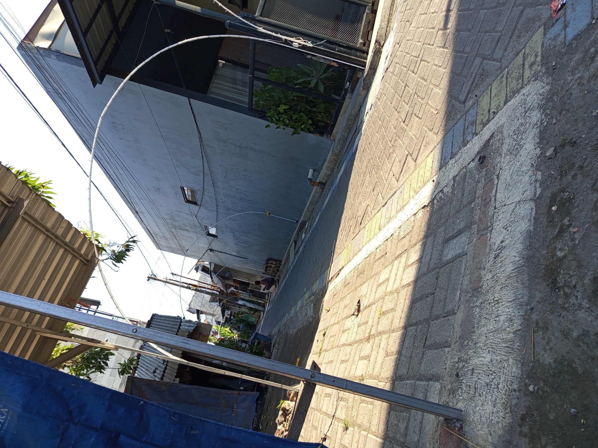 In the city of Tebel, Indonesia, an image captures a narrow, paved alleyway lined with buildings. The alley is predominantly constructed with interlocking brick pavers, with a painted white and yellow line marking the edge of the walkway. On the left, a tall, dark blue tarp is draped over a metal structure, partially obscuring a glimpse of buildings and foliage behind it. A person is seen squatting in the distance on the right side of the alley, near a stack of wooden pallets. Overhead, a complex network of wires crisscrosses the sky, supported by utility poles. The buildings flanking the alley exhibit a mix of architectural styles, with one on the right featuring a modern, angled design with a grey exterior and dark-framed windows. Green foliage is visible in a fenced-off area on the right, adding a touch of nature to the urban setting. The sunlight casts distinct shadows across the alley, indicating it is a bright day.