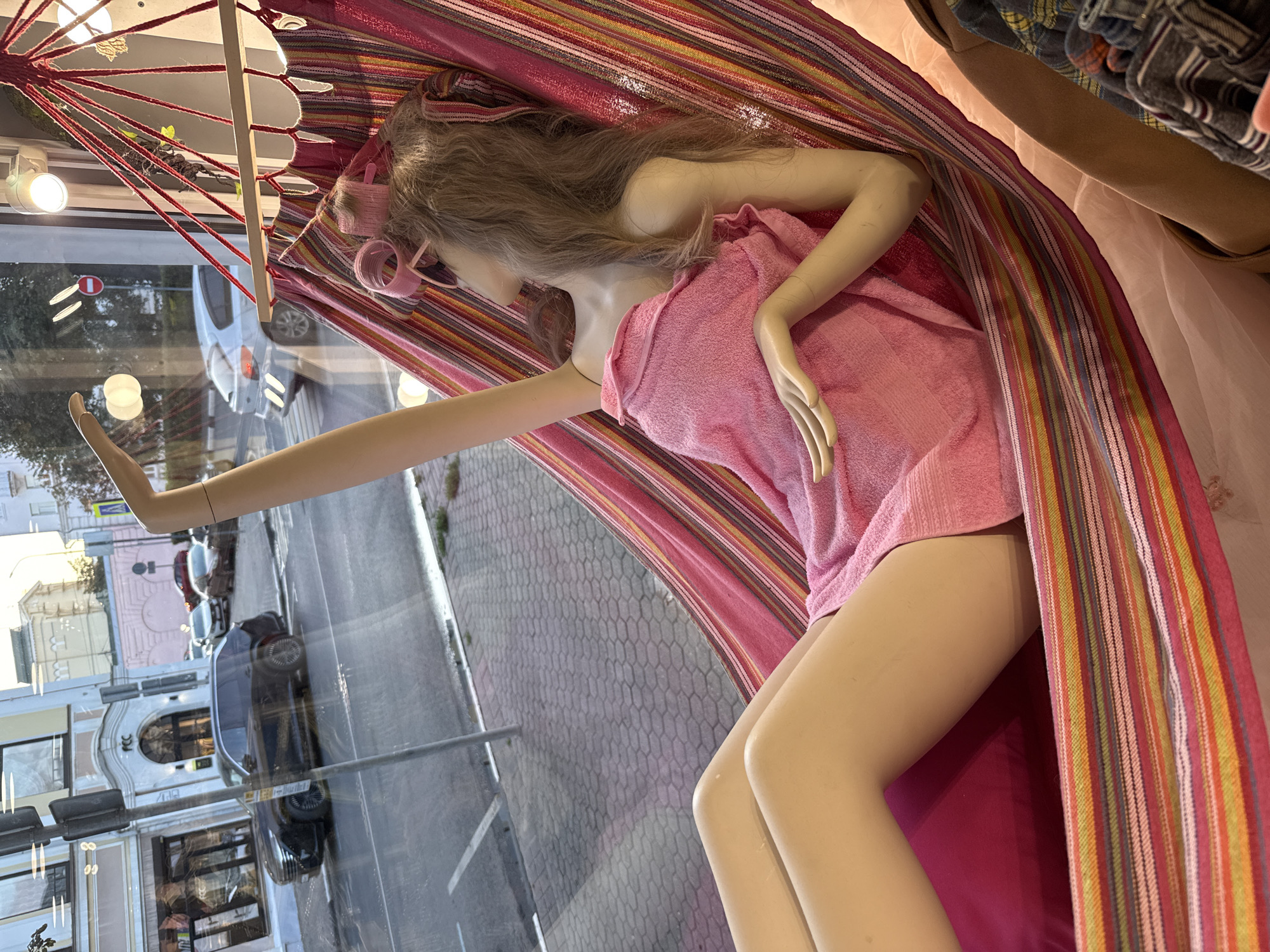 A mannequin, dressed in a pink towel and with curlers in its hair, is posed in a hammock. The hammock has a striped pattern of pink, red, yellow, and green. The mannequin's right arm is extended, and its left arm is bent at the elbow. The scene is viewed through a window, which reflects a street scene with cars and buildings. The sky appears overcast, suggesting it might be daytime. The overall impression is that of a display, possibly in a store window, designed to evoke a sense of relaxation or a spa-like atmosphere. There is no visible text in the image.