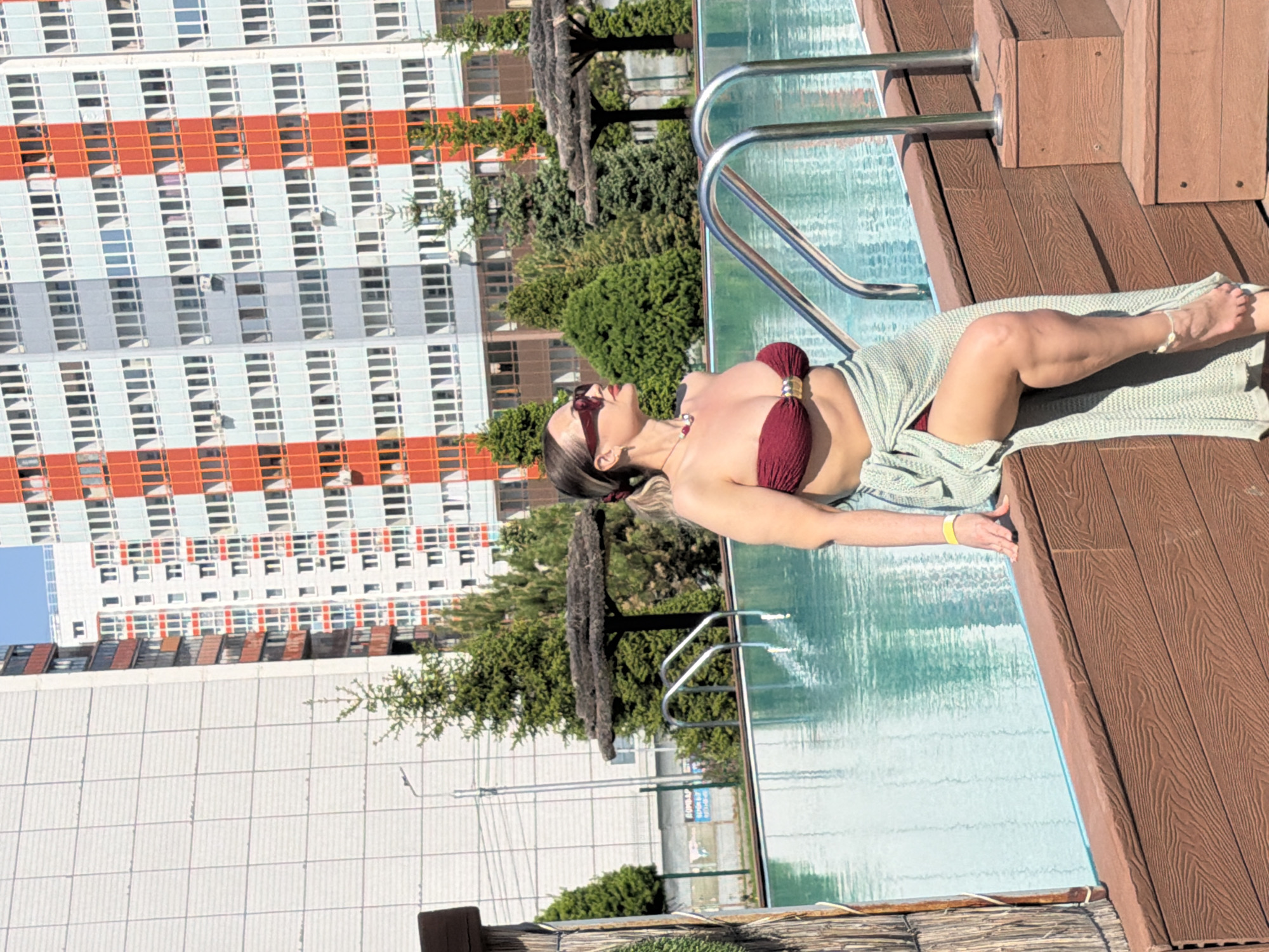 An outdoor scene captured on a sunny day features a woman relaxing by a swimming pool, likely in Krasnoyarsk, Russia. The woman, appearing to be in her 20s or 30s, is lying on her back on a brown wooden deck adjacent to the pool. She wears a dark red bikini top with a gold-colored embellishment and dark sunglasses, with a light green, textured wrap or towel draped over her legs. Her dark hair is pulled back, and she has a necklace. Her left leg is slightly bent, and an anklet is visible on her right foot.
The swimming pool contains clear, light blue water, reflecting the bright sky. A silver metal pool ladder provides access to the water. In the background, a tall, imposing residential building stands prominently, characterized by numerous windows arranged in a grid pattern and distinct horizontal bands of light grey, white, and orange-red sections. Green trees and bushes are visible between the pool area and the building, adding natural elements to the urban setting. The strong natural light indicates a clear, sunny day, with visible shadows on the deck. The overall ambiance suggests a moment of leisure in a city environment. A blurry, illegible sign can be seen in the distant background.