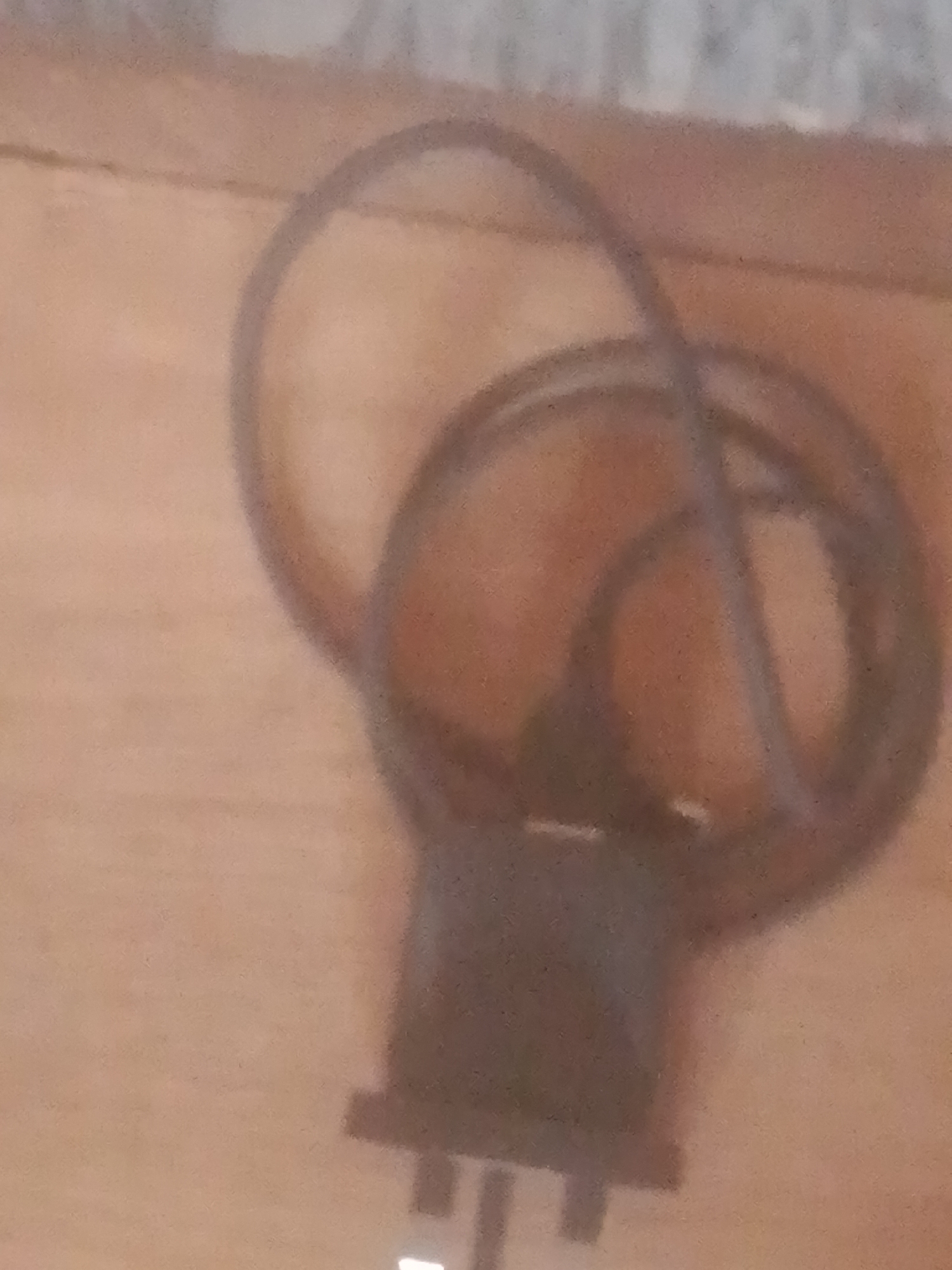 This is a close-up, blurry photograph of a dark, coiled electrical cord with a plug. The cord is wrapped around itself, forming a rough oval shape, with the plug at the bottom. The plug appears to have three prongs, suggesting it is a Type G plug, commonly used in the UK and other countries. The cord and plug are resting on a wooden surface, which has a visible grain pattern. The background is out of focus and appears to be a textured surface, possibly a wall or fabric. There are no people visible in the image, nor any clear indication of the time of day, weather, or specific location beyond the general context of Nigeria. The image is dimly lit, and the blurriness makes it difficult to discern finer details. The overall impression is of a common household or electronic item.