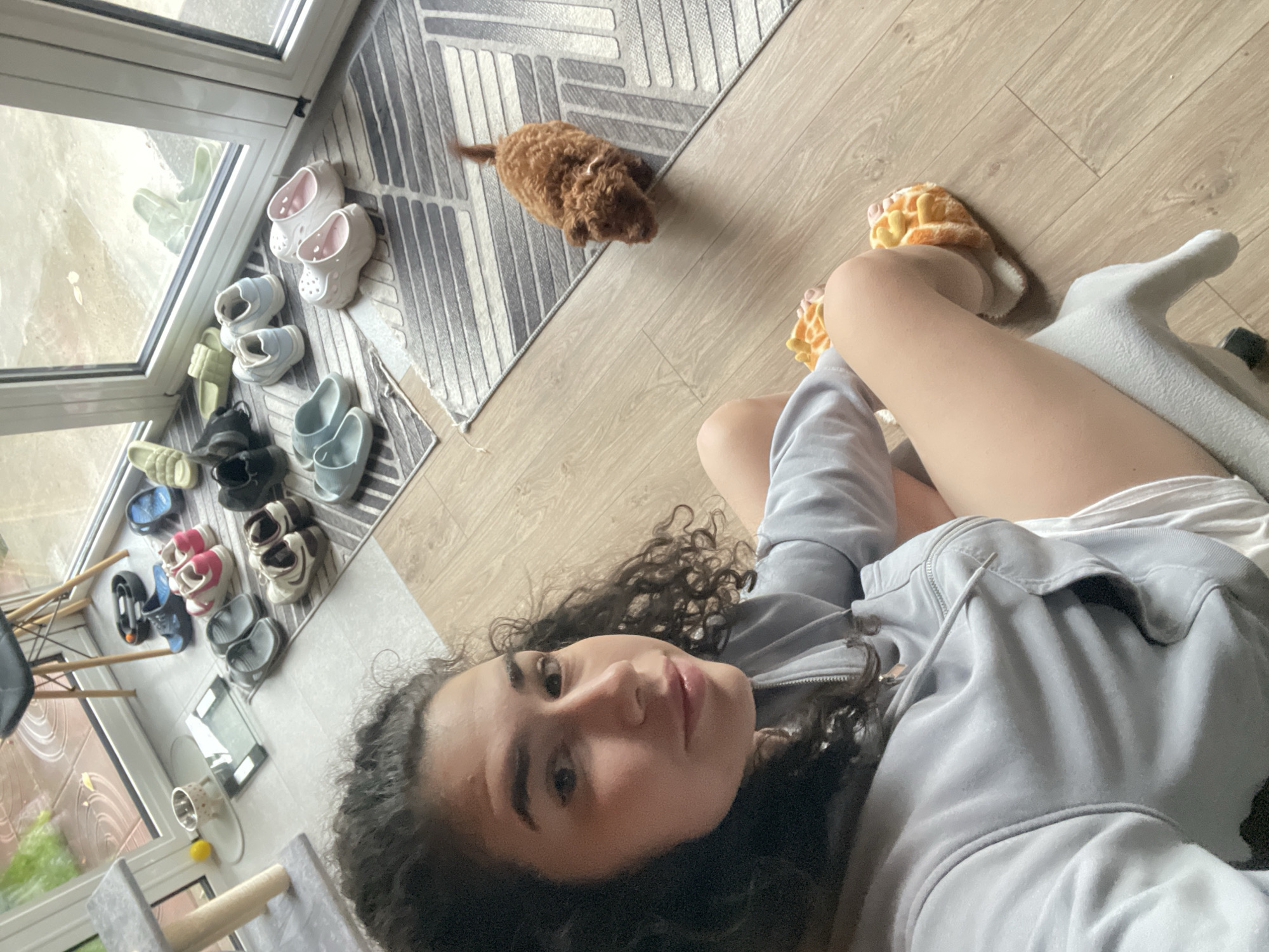 A young woman with curly dark hair, wearing a grey hooded sweatshirt and white shorts, is sitting on the floor in what appears to be a modern apartment or house, possibly in Sofia, Bulgaria, given the context. She is looking at the camera and has her legs crossed. She is wearing fluffy, waffle-patterned slippers. To her right, a small, light brown poodle puppy is walking on a grey and white striped rug. The background shows a large glass door leading outside to a tiled patio with some greenery visible. Several pairs of shoes, including sandals and sneakers, are neatly arranged on the floor and on mats near the door. A digital scale and a cat tree are also present in the room. The flooring is light wood laminate.