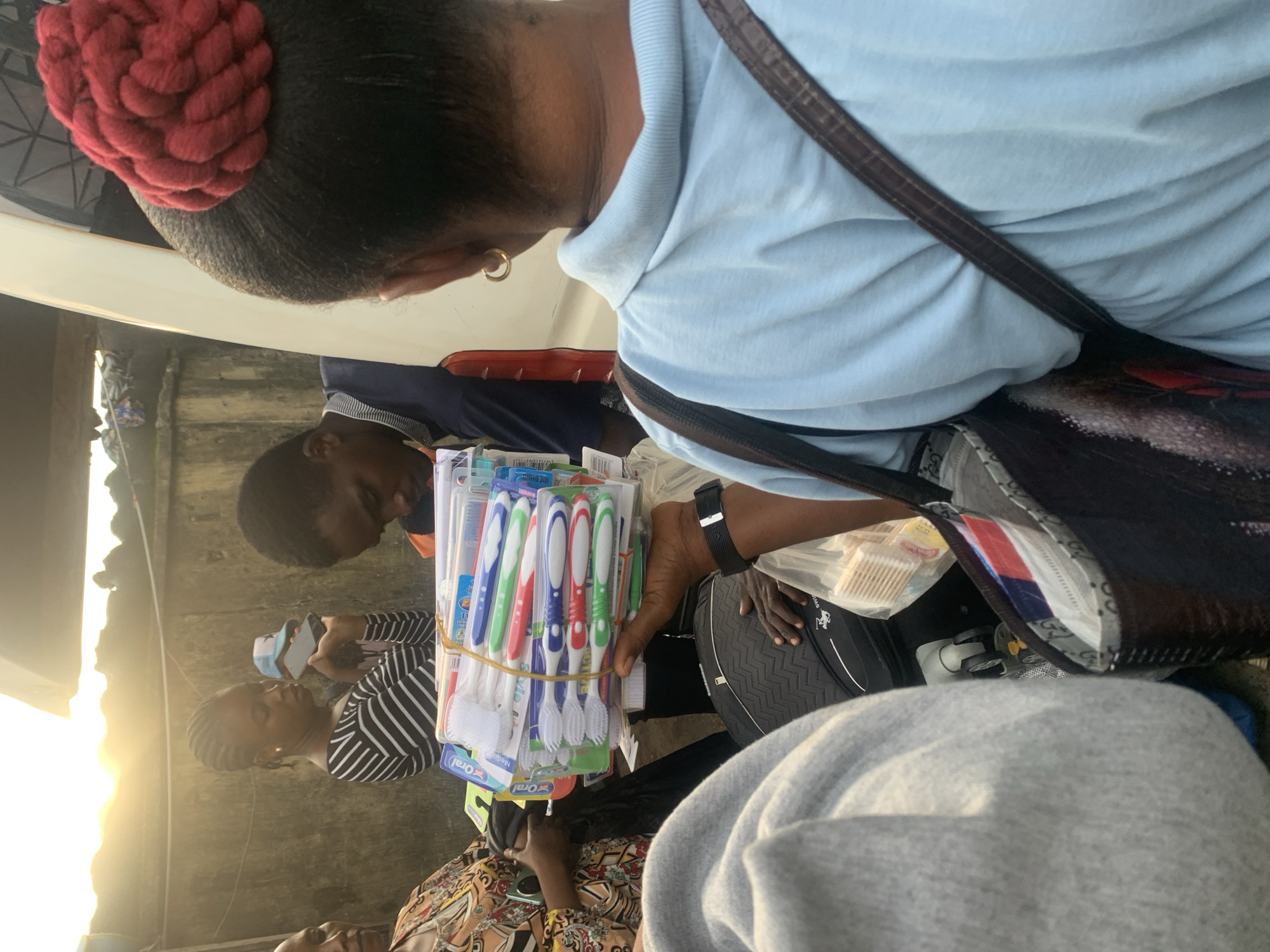 In Elelenwo, Nigeria, an outdoor scene depicts a group of people near a vehicle. A young woman with braided hair, wearing a black and white striped shirt, holds a smartphone and a blue and white cap. Beside her, a young man in a blue and orange shirt looks down. In the foreground, a person in a light blue shirt with a red braided hairstyle tied up on their head holds a stack of toothbrushes packaged in plastic. They are wearing a gold hoop earring and a dark brown crossbody bag. Another individual, seen from the back, wears a grey hooded garment. A large dark bag with the brand name "SWISS" visible is on the ground. The lighting suggests it is late afternoon or early morning, with the sun low in the sky. The background consists of a textured wall and a portion of a white vehicle.