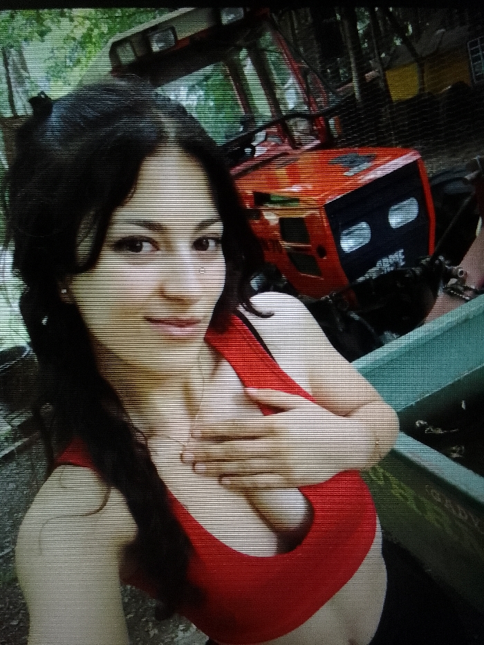 The image displays a close-up portrait of a female individual in an outdoor, likely rural or semi-rural, setting. The location is in the city of Unknown, Unknown.
The subject is a young adult female with long, dark, wavy hair, dark eyes, and a light-to-medium skin tone. She has a small stud piercing in her left nostril. She is wearing a red sports bra or crop top, and a thin necklace with a small pendant is visible on her chest. Her right hand is positioned over her upper chest/décolletage area. She is looking directly at the camera with a subtle smile.
In the background, a bright orange and black tractor is partially visible behind and to the right of the individual. Its front section, including the black grille and two square headlights, is discernible. Green foliage and trees are visible further in the background, suggesting an outdoor environment. To the lower right of the individual, a segment of a teal or green structure, possibly a trough or a part of other machinery, is present. The entire image is overlaid with prominent horizontal scan lines, indicative of being a photograph of a screen or a specific digital capture process.